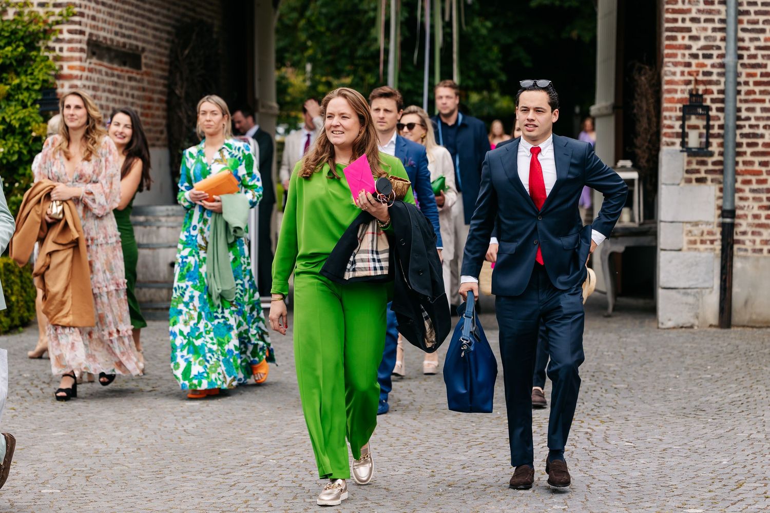 Wedding guests in vibrant outfits, including a green dress and navy suit, walk along a cobblestone path.
