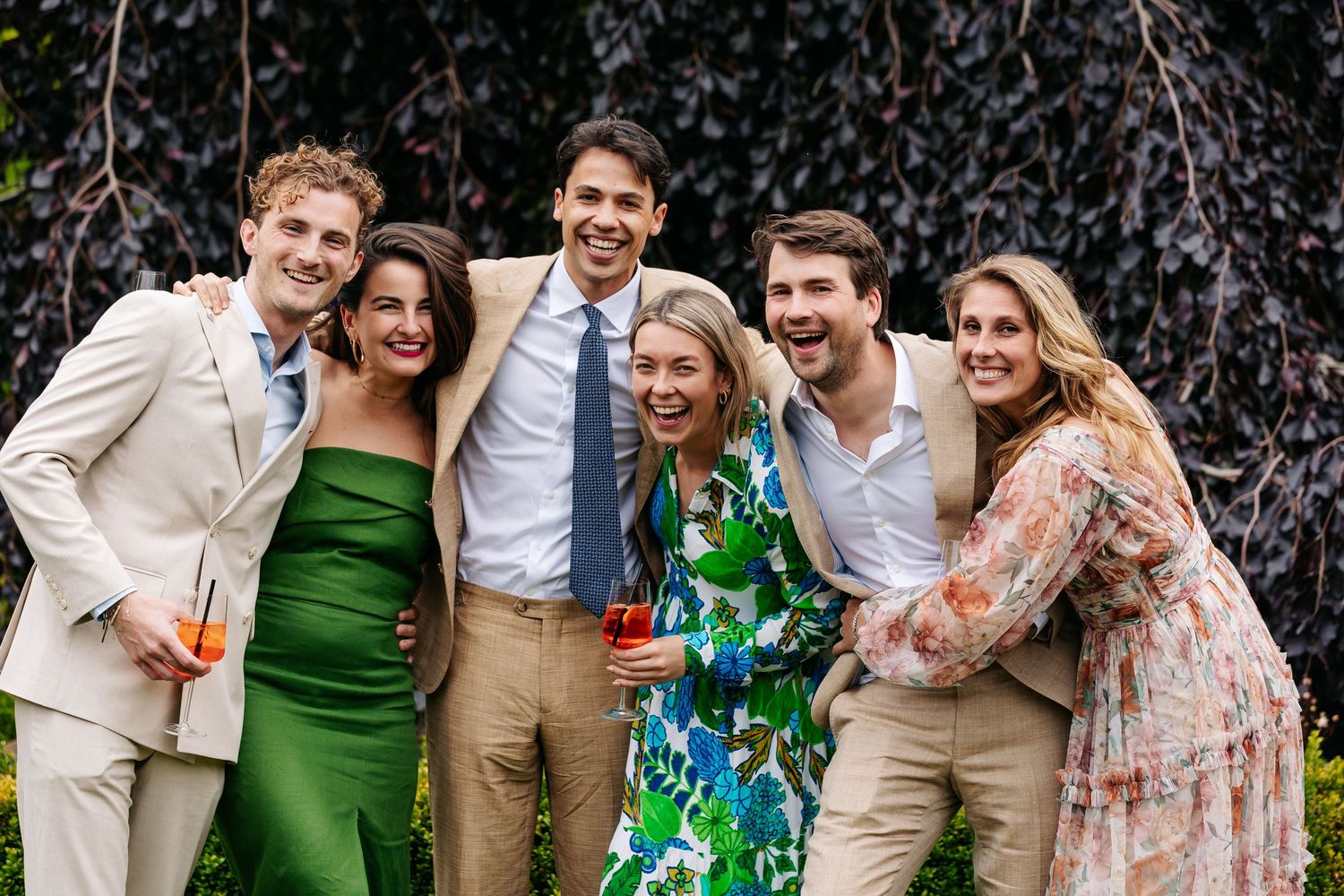Six young wedding guests laughing and posing together outdoors in colorful attire.