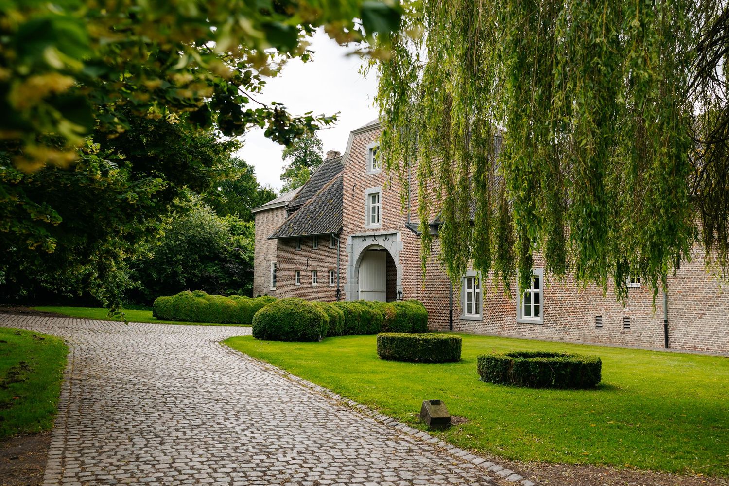 Historic brick manor house with manicured hedges and cobblestone driveway framed by weeping willow branches.