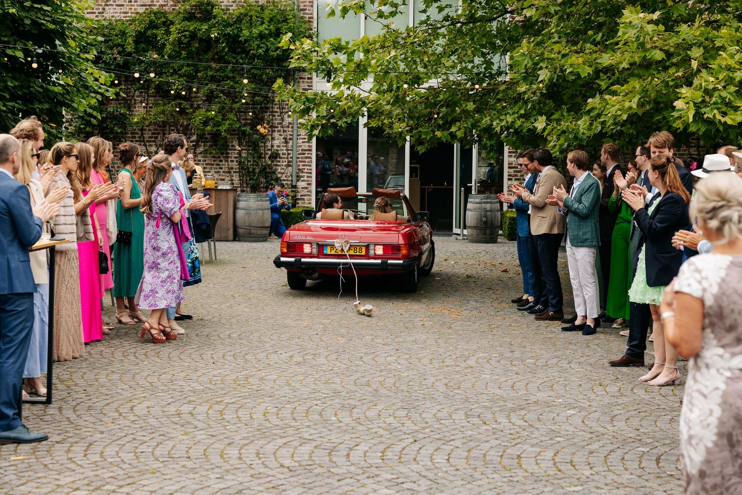 Wedding guests lining a cobblestone courtyard applauding a red vintage convertible car.