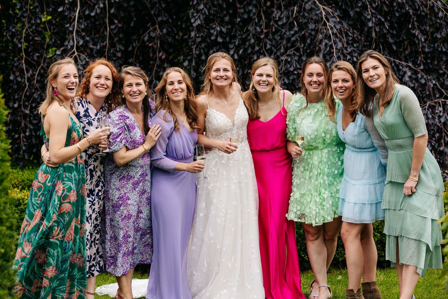A bride in a white lace gown poses with eight smiling bridesmaids in colorful dresses outdoors.