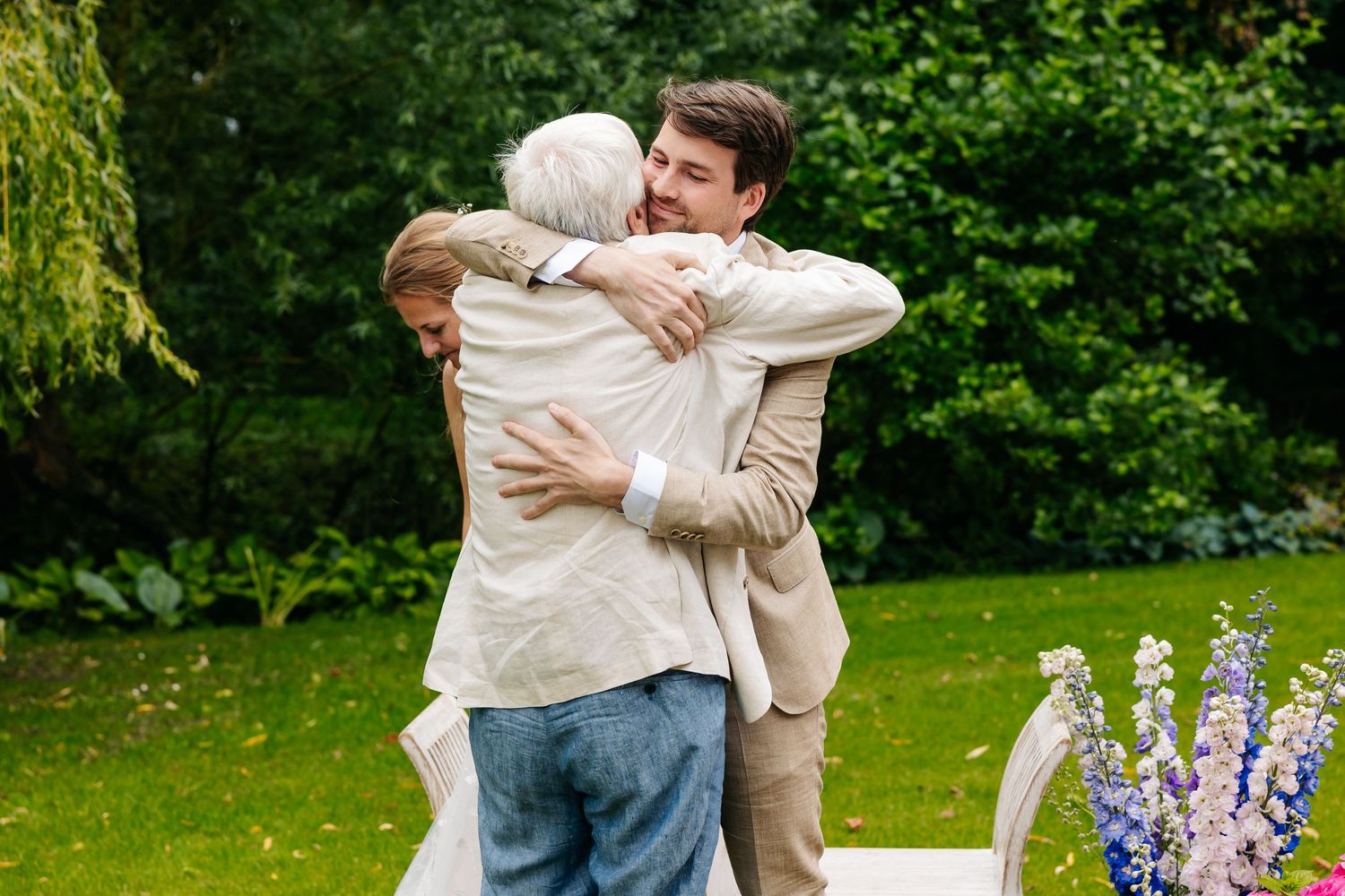 Groom in a beige suit shares a warm embrace with an elderly guest in a lush garden setting.