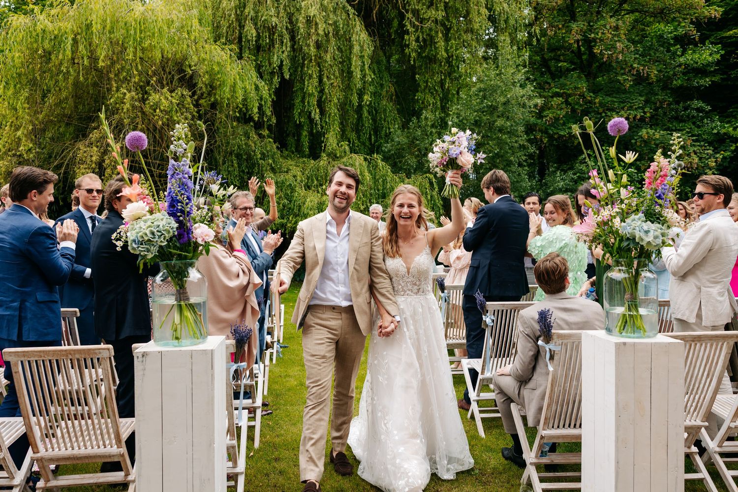 Newlyweds walk joyfully down outdoor aisle as guests cheer at garden wedding ceremony.