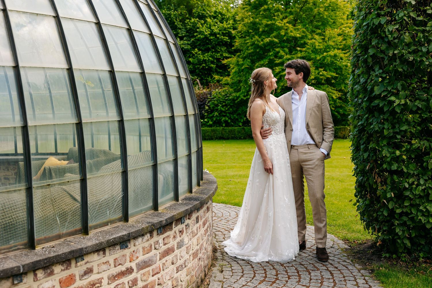 Bride and groom sharing a romantic moment beside a domed glass greenhouse surrounded by lush green gardens.