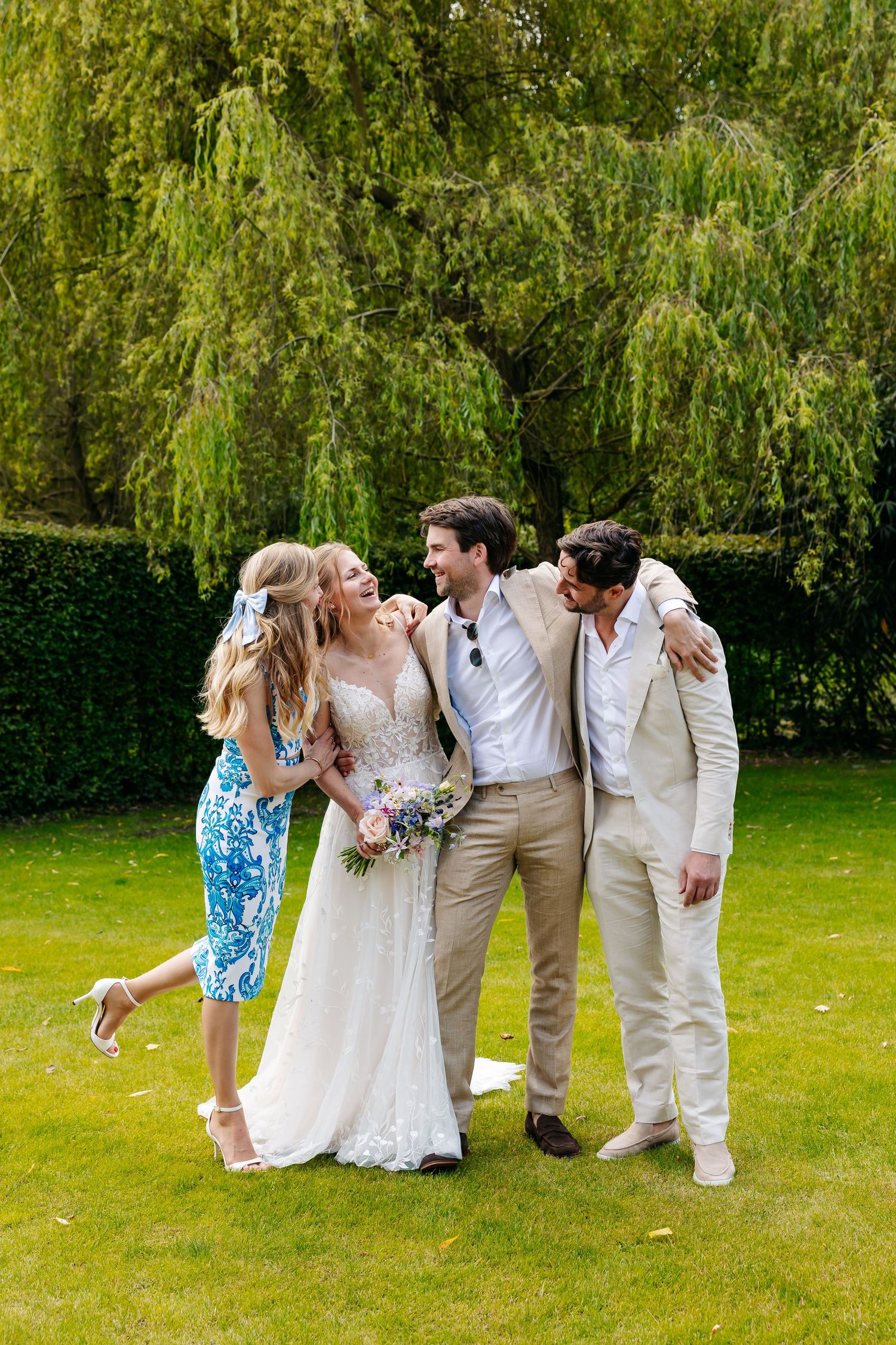Bride and groom laughing with two guests on green lawn in front of weeping willow tree.