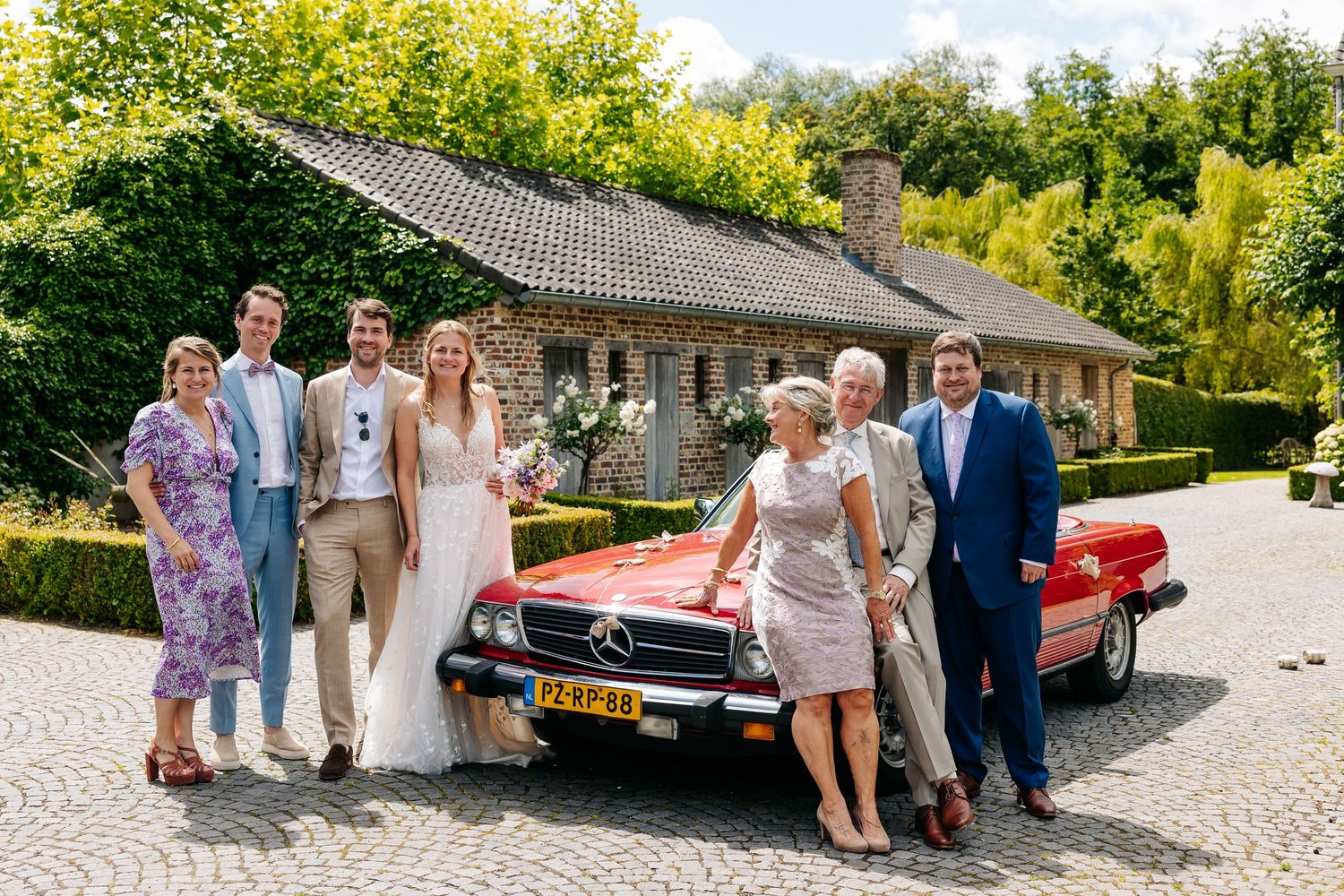 Wedding party posing with vintage red Mercedes-Benz convertible on cobblestone driveway outdoors.