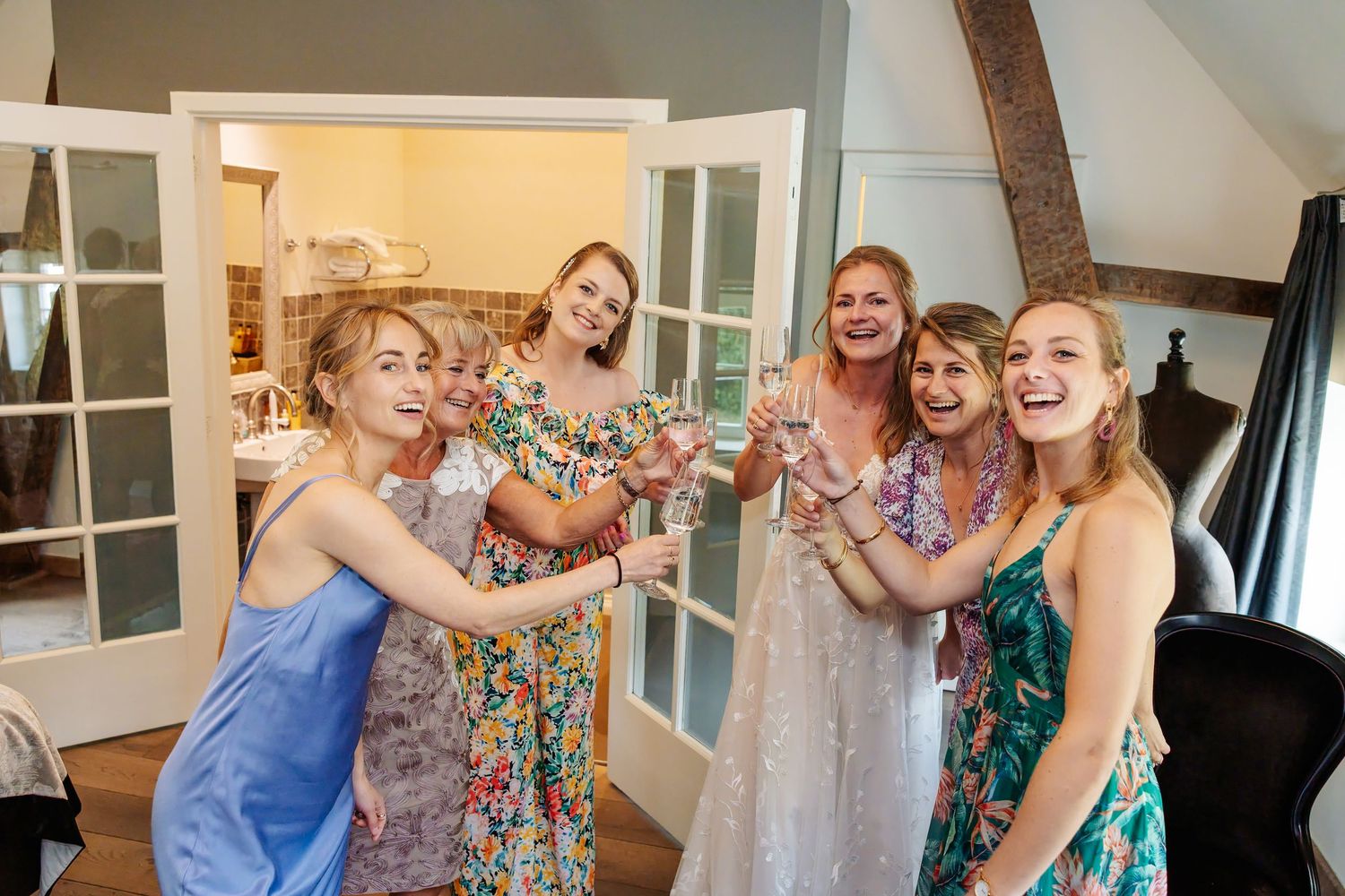 Bride and bridesmaids laughing and toasting with champagne glasses in rustic bridal suite.