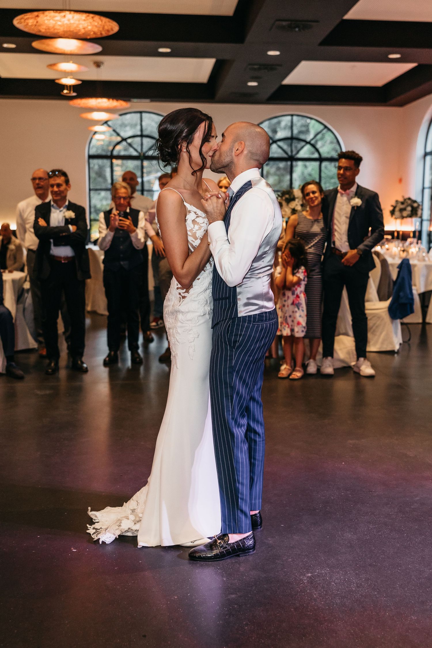 Bride and groom sharing romantic kiss during first dance at wedding reception with guests applauding around them.