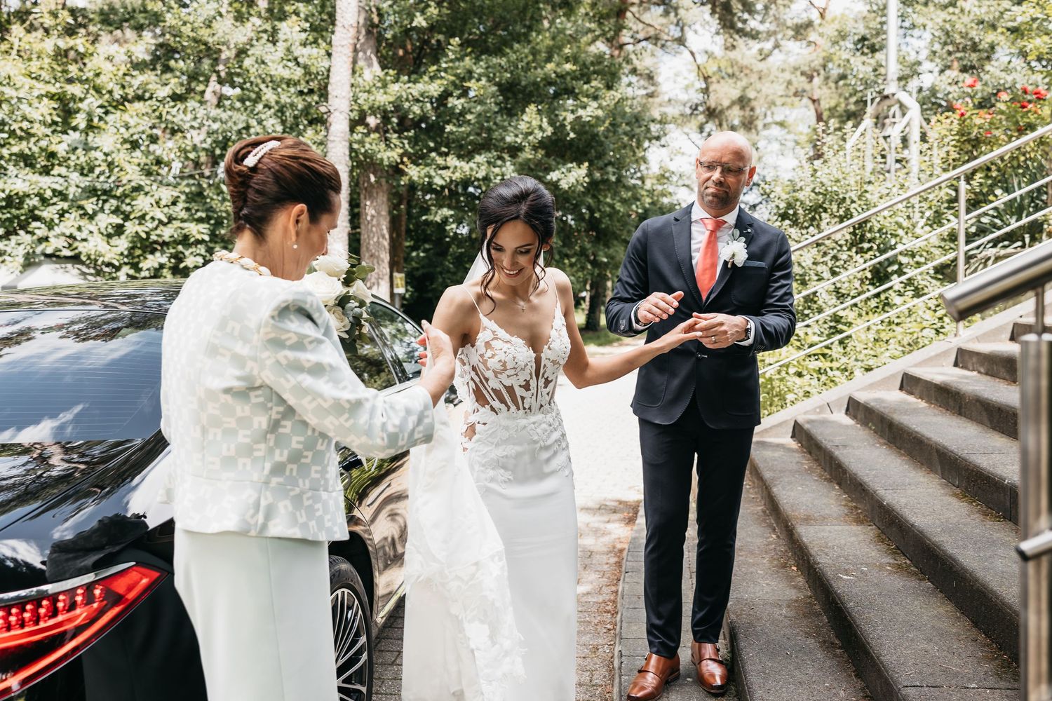 Bride in lace wedding dress assisted by family outside church before ceremony.