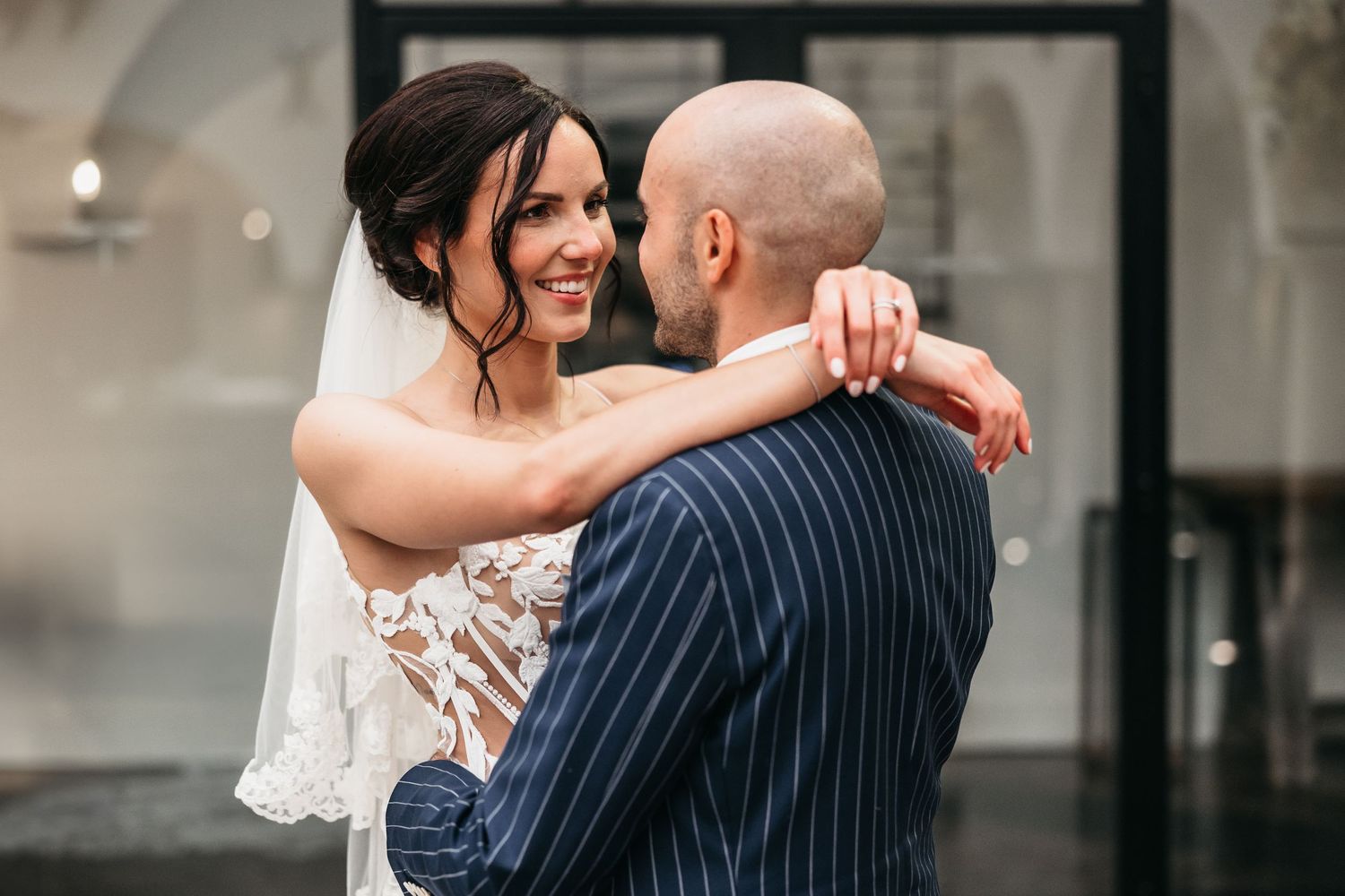 Smiling bride in lace wedding gown embraces bald groom in navy pinstripe suit outdoors.
