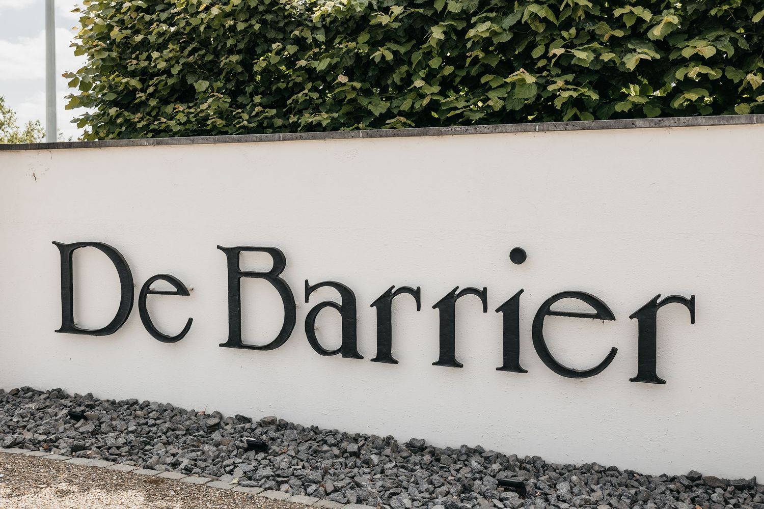 Elegant De Barrier wedding venue sign mounted on white wall surrounded by green hedges.