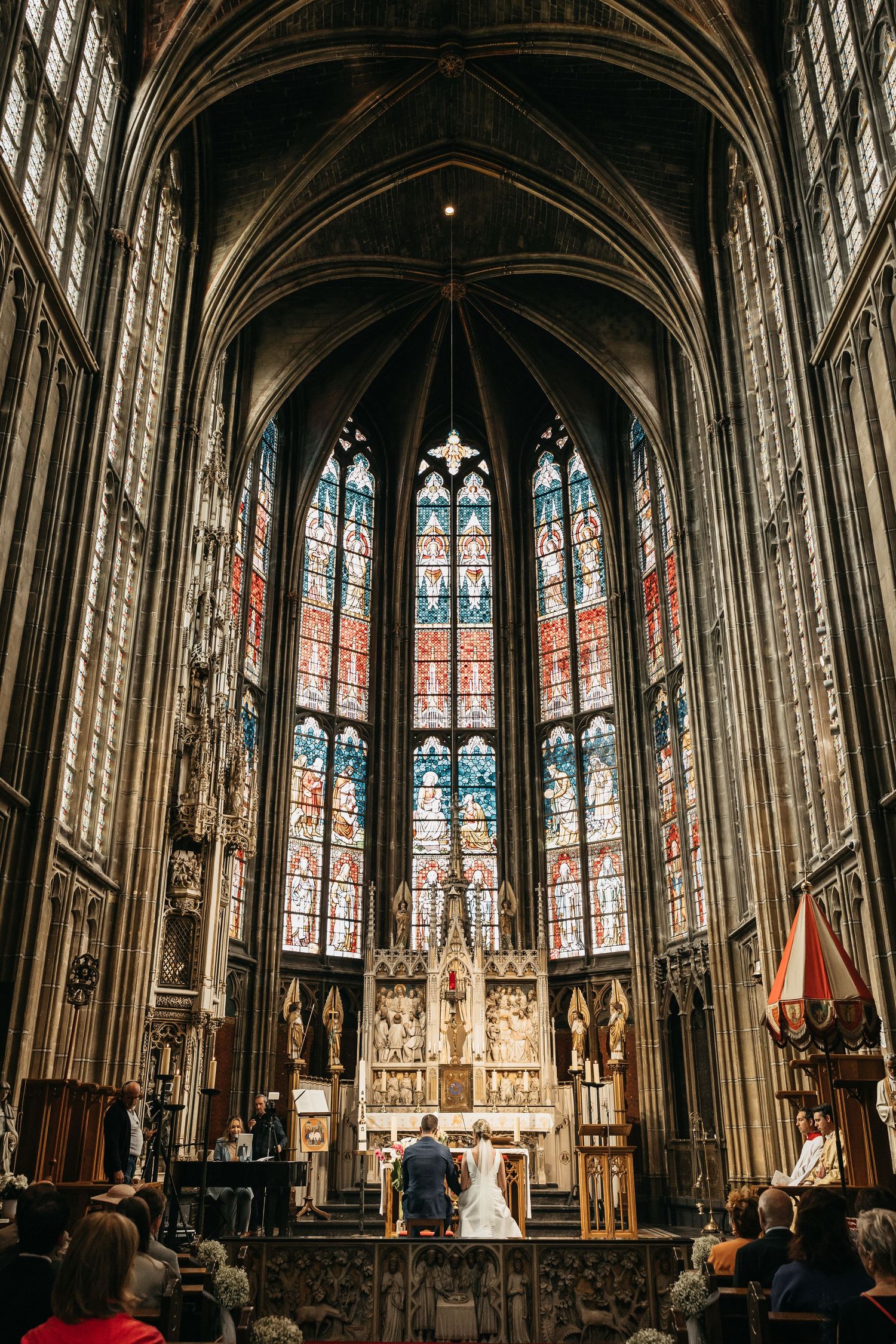 Bride and groom stand at ornate altar beneath stunning stained glass windows in Gothic cathedral.