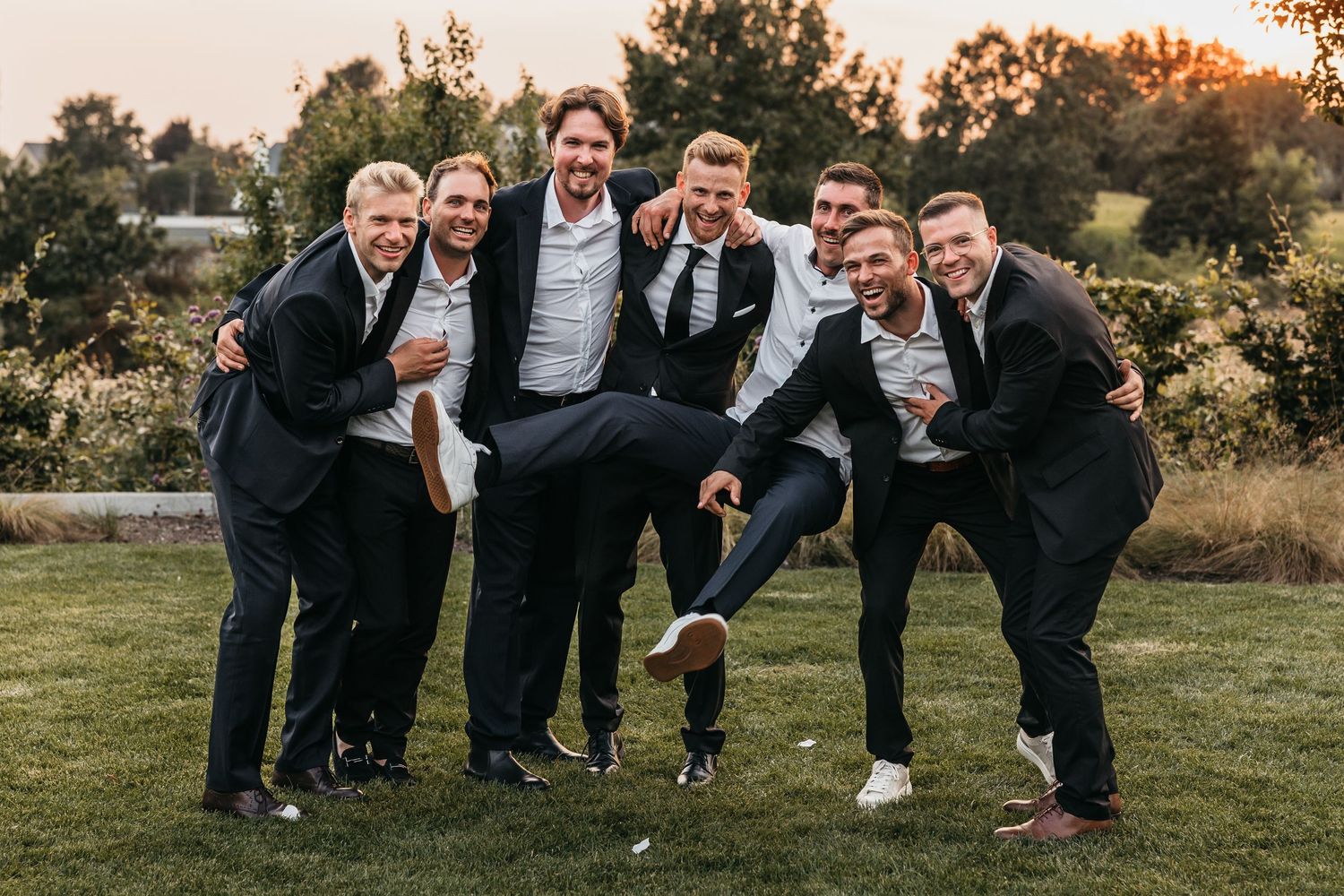 Groomsmen in dark suits laughing and posing playfully together on grass at wedding.