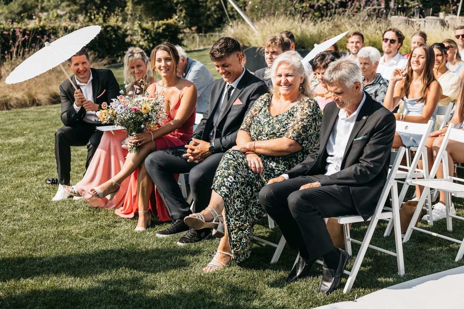 Wedding guests smiling and laughing while seated in white chairs during outdoor garden ceremony.
