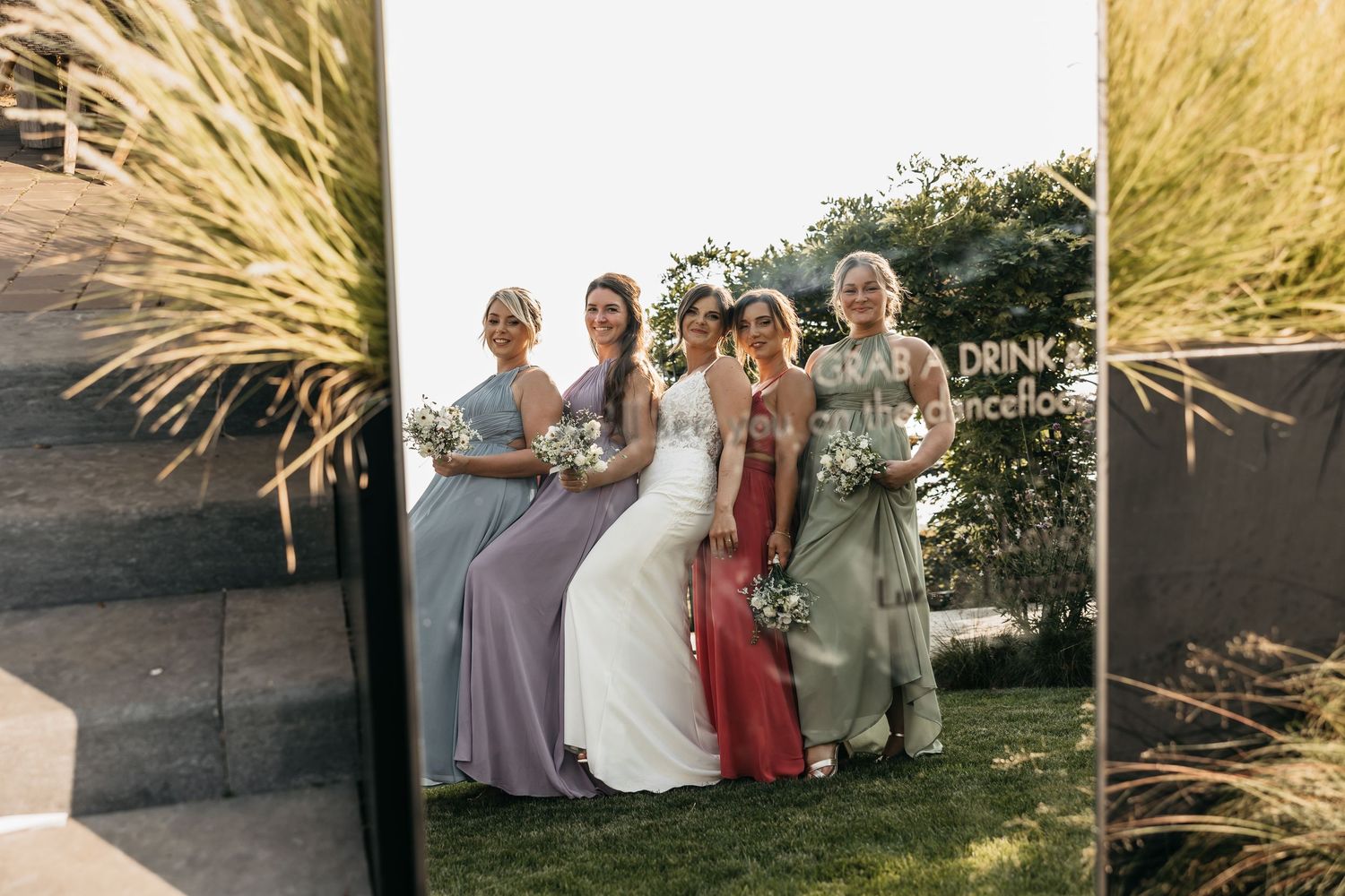 Bride and bridesmaids in pastel gowns smiling together reflected in a mirror at outdoor wedding venue.