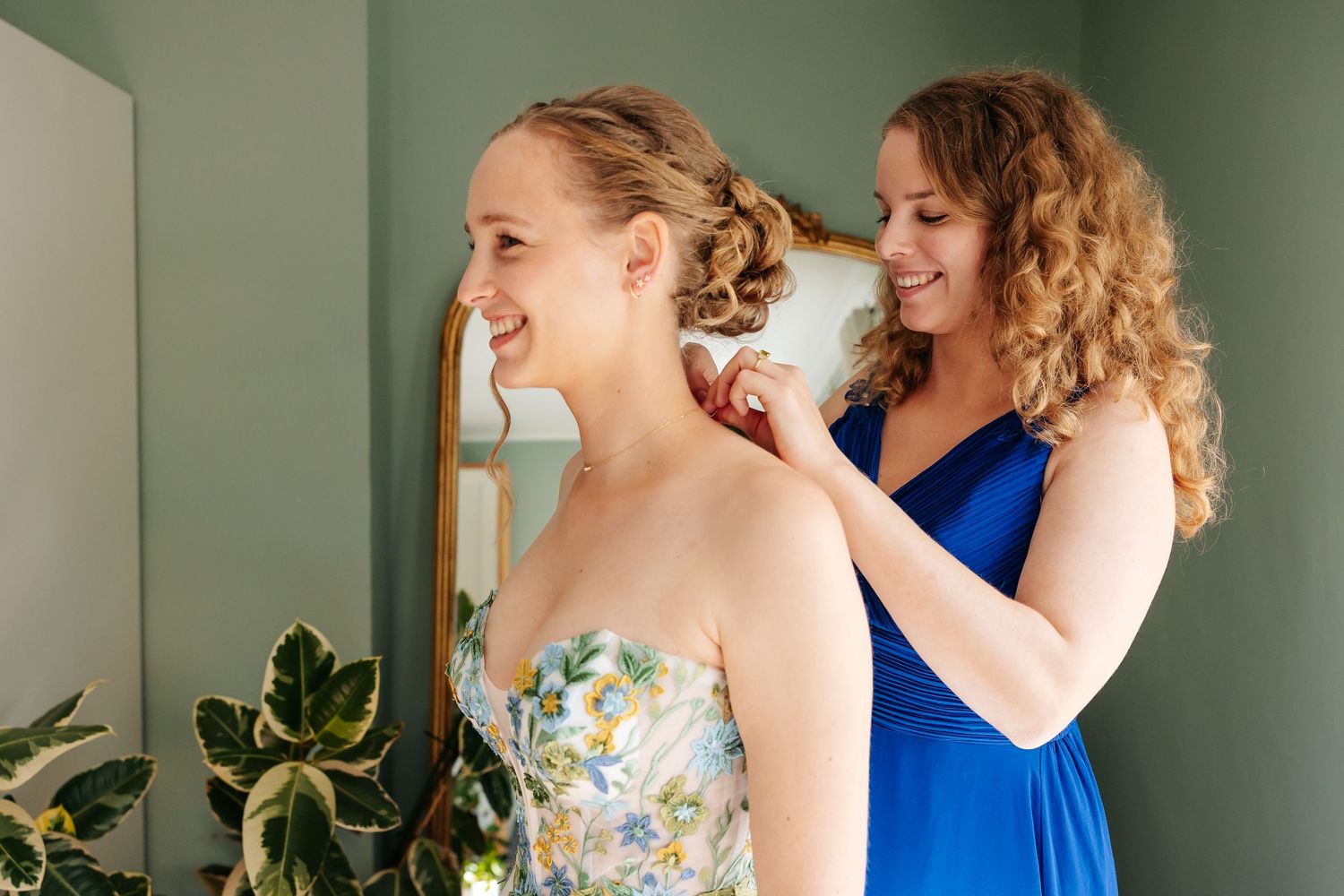 Bridesmaid in blue dress helping bride with earring while bride smiles in floral wedding gown.