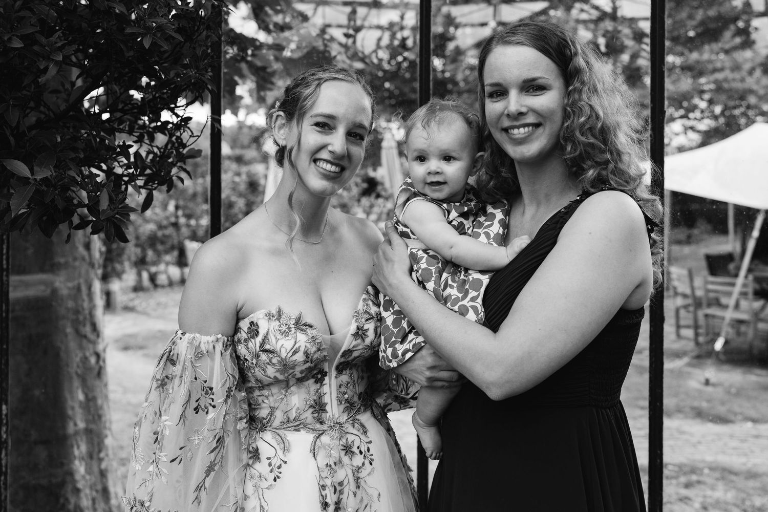 Black and white photo of a bride, bridesmaid, and baby smiling together at a wedding.