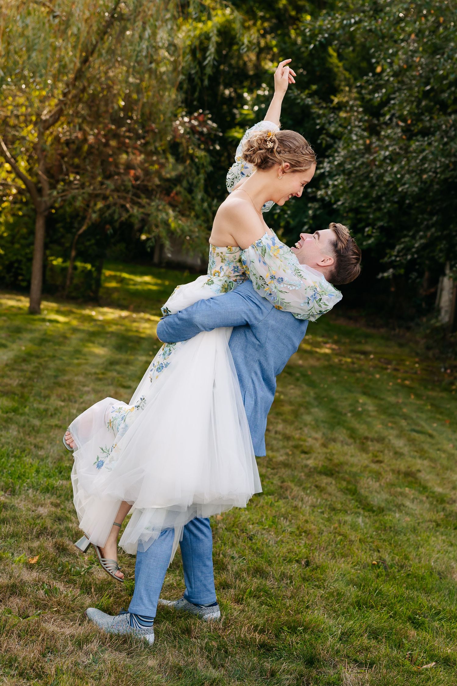 Groom lifts bride joyfully in a lush garden, bride in floral dress raises her arm gracefully.