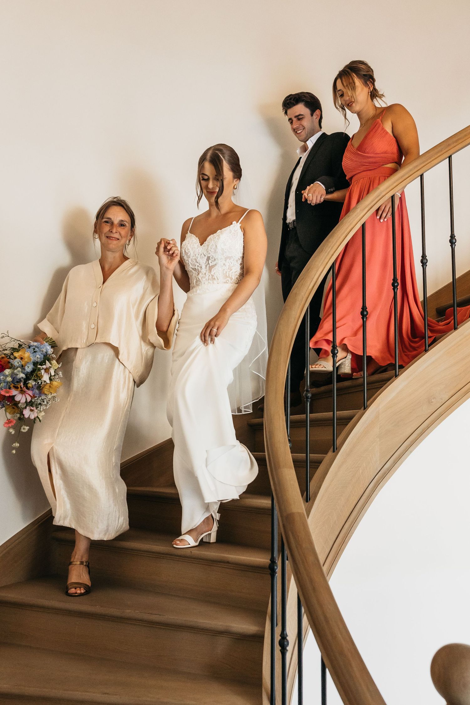Bride in white gown descends curved staircase with family members in coral and cream outfits.