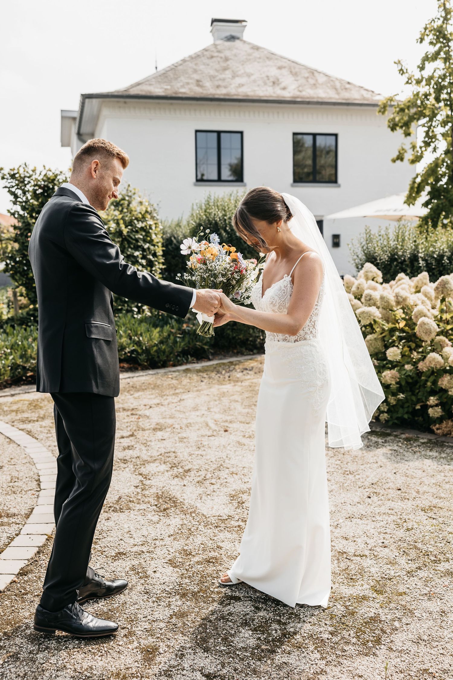 Groom holds bride's hand while she looks down at bouquet in sunlit garden.