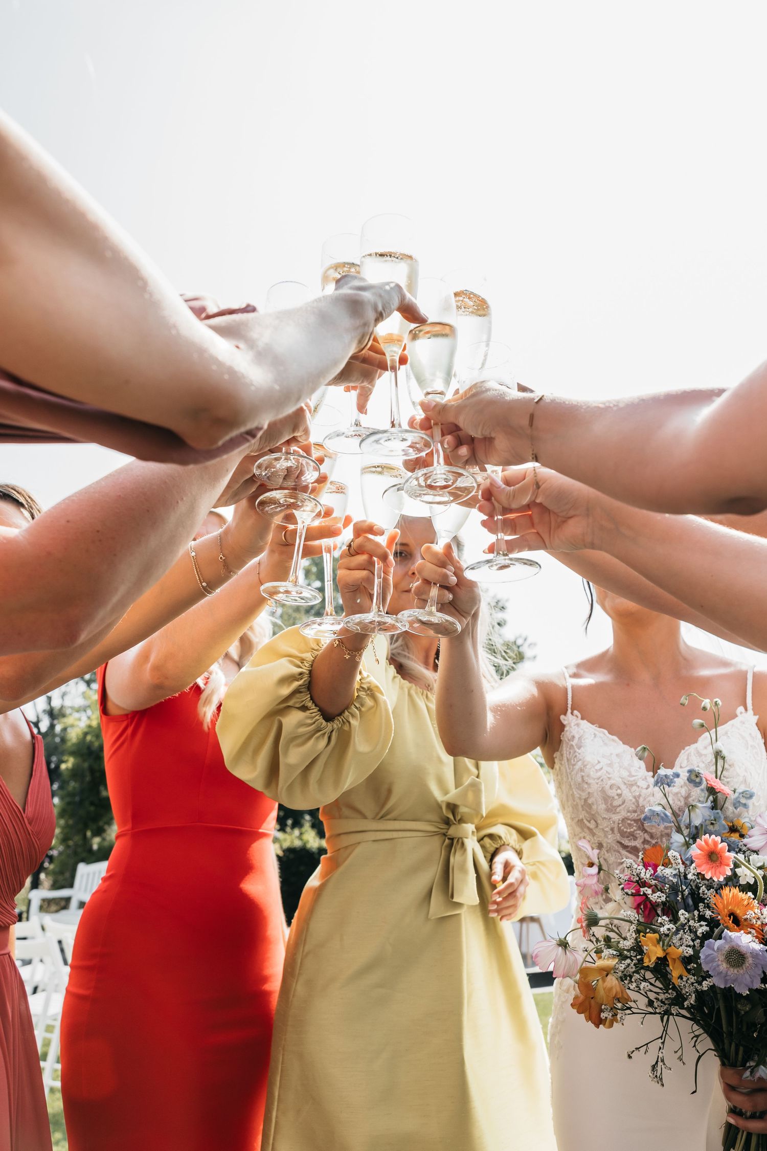 Wedding party clinks champagne glasses in celebratory toast at outdoor wedding with bride holding floral bouquet.