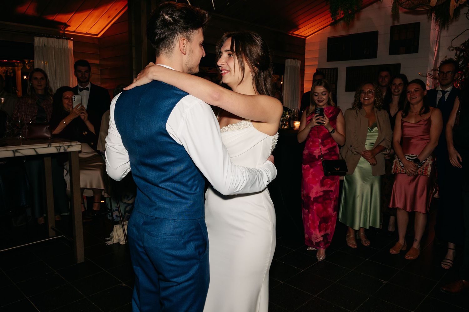 Bride and groom share their first dance, embracing closely while wedding guests watch and celebrate in the background.