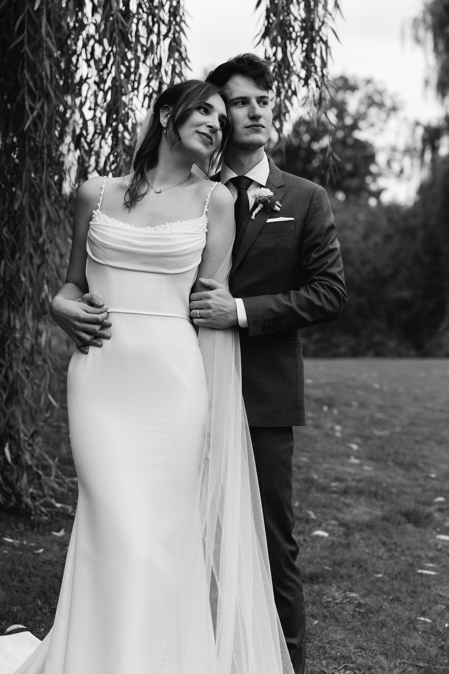 Black and white portrait of bride and groom embracing tenderly under weeping willow tree outdoors.