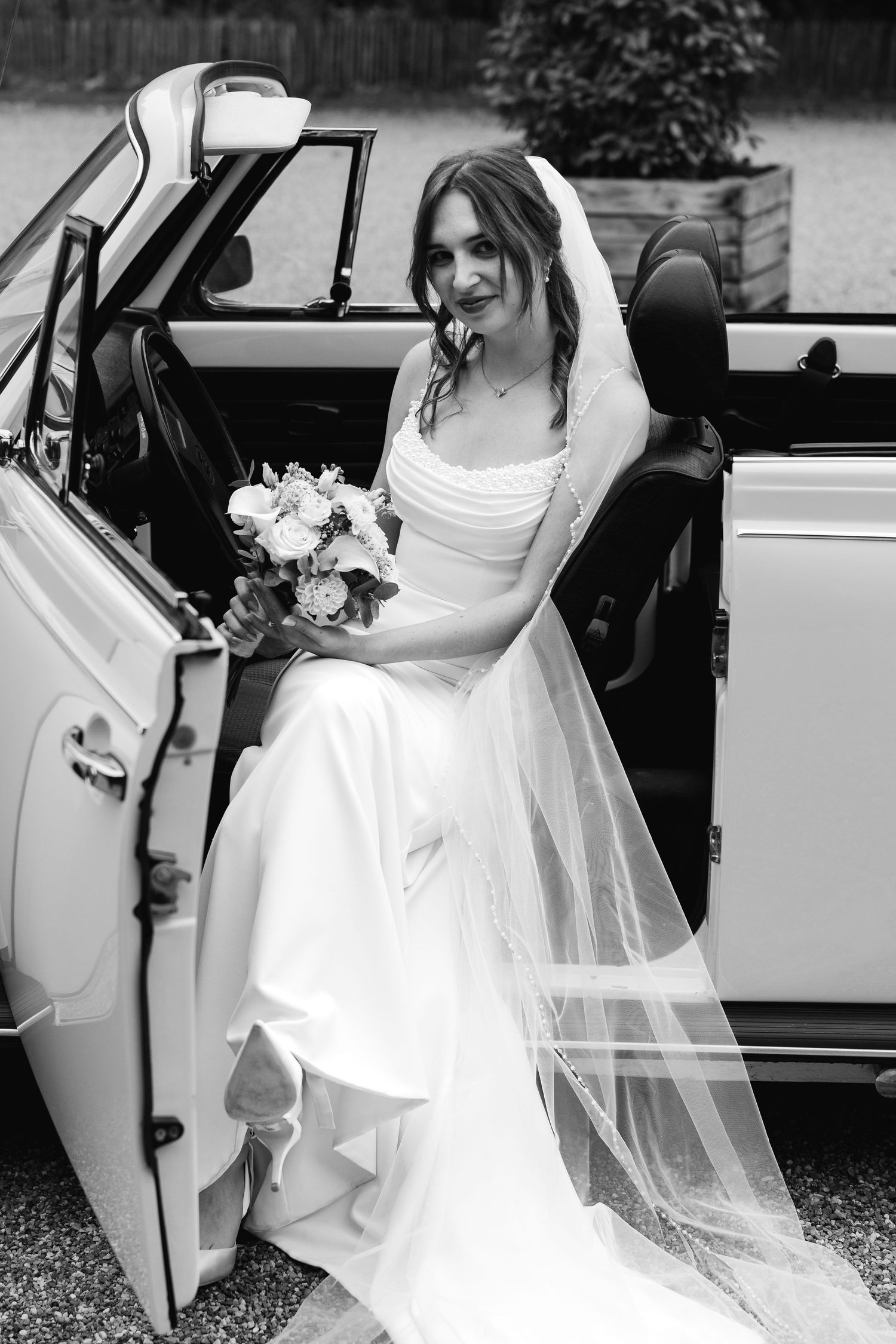 Black and white photo of bride in white gown and veil sitting in classic white convertible car.