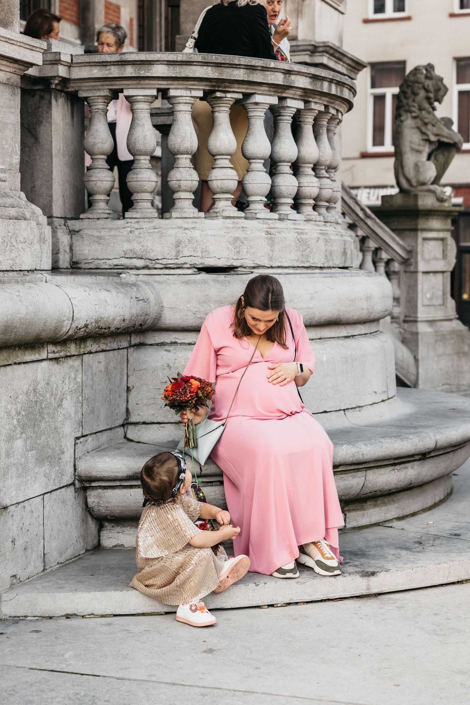 Pregnant woman in pink dress sits on stone steps with young child holding red floral bouquet.