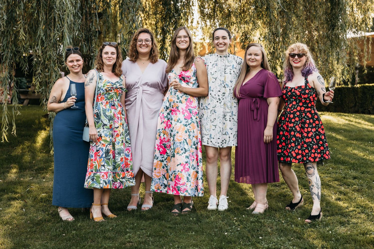 Seven female wedding guests smiling together in colorful summer dresses under a willow tree.