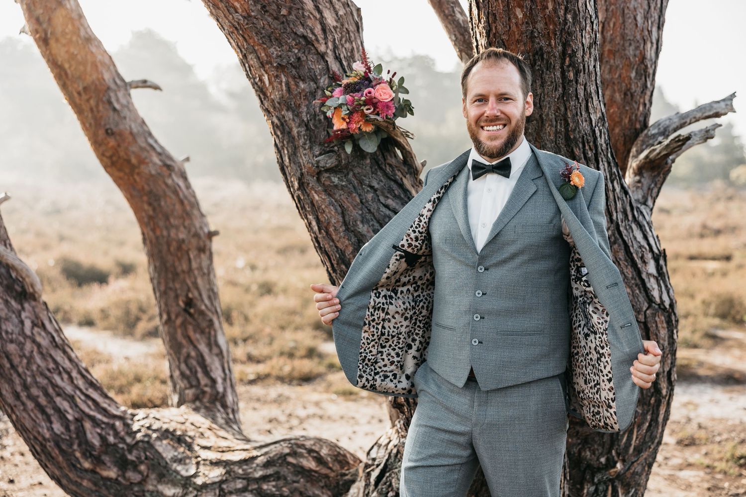 Smiling groom in grey suit opens jacket to reveal leopard print lining while posing against a pine tree.