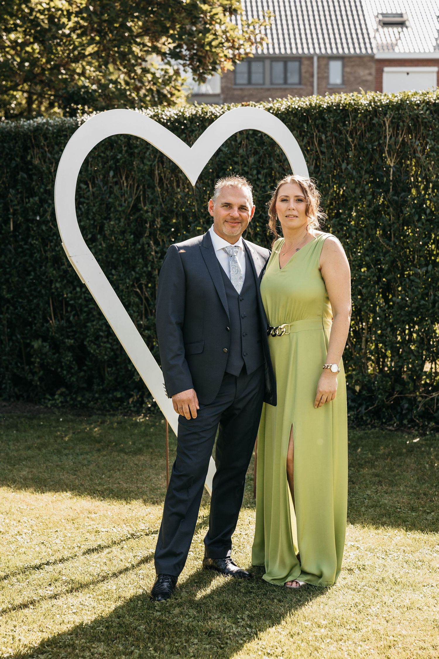 Elegant couple in formal attire poses in front of a large white heart decoration in a garden.