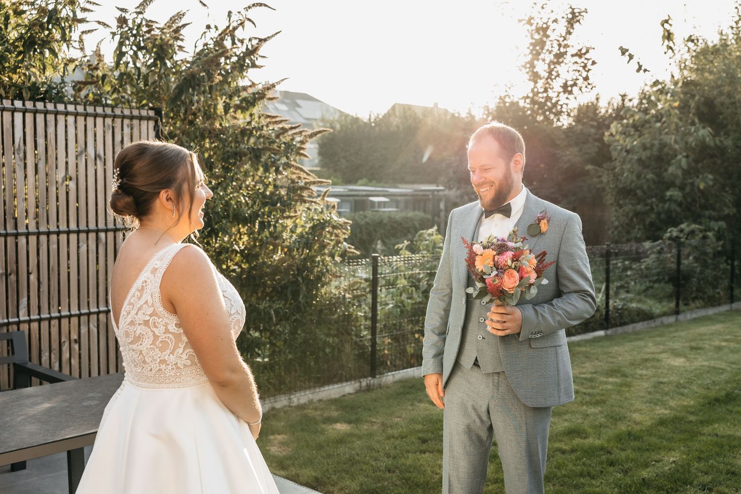 Groom in gray suit smiles holding bridal bouquet as bride in lace gown approaches for first look at sunset.