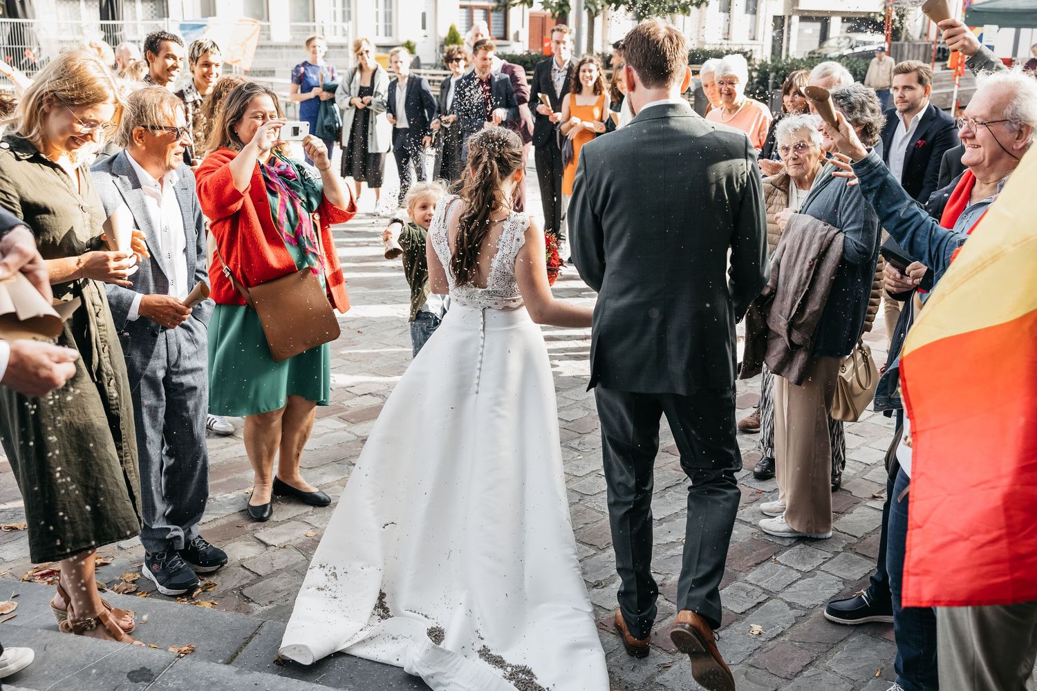 Bride in white gown and groom greet guests in sunny outdoor plaza after wedding ceremony.