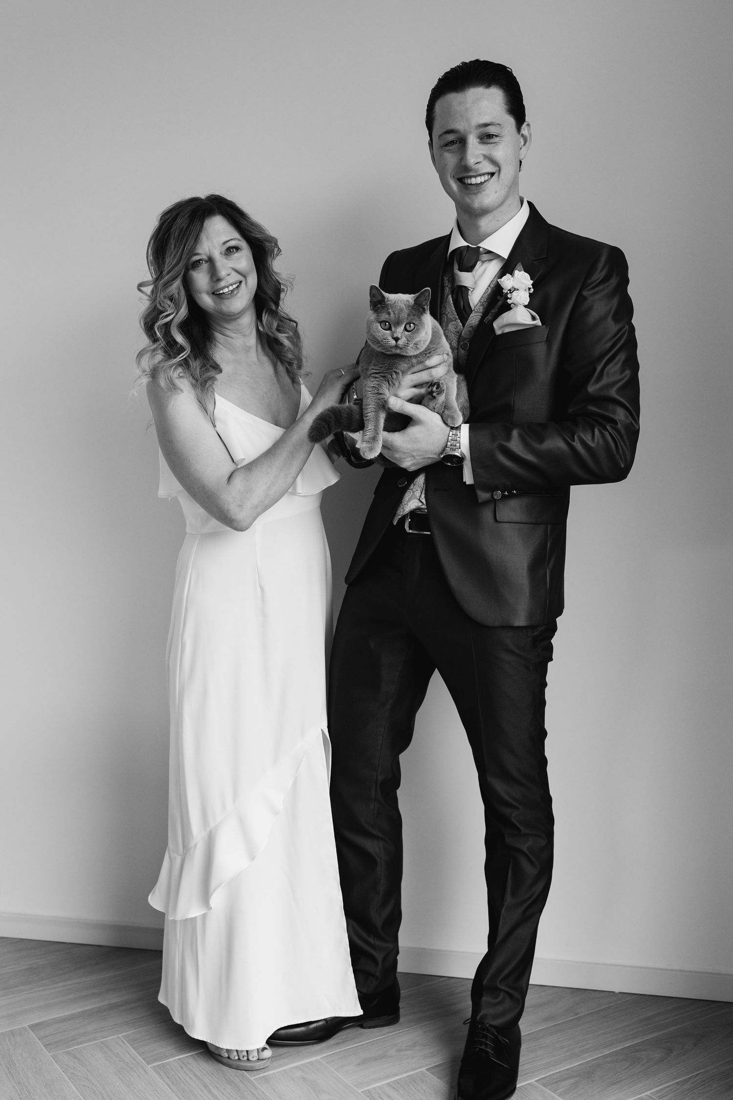 Black and white photo of bride and groom in wedding attire smiling while holding their gray cat.