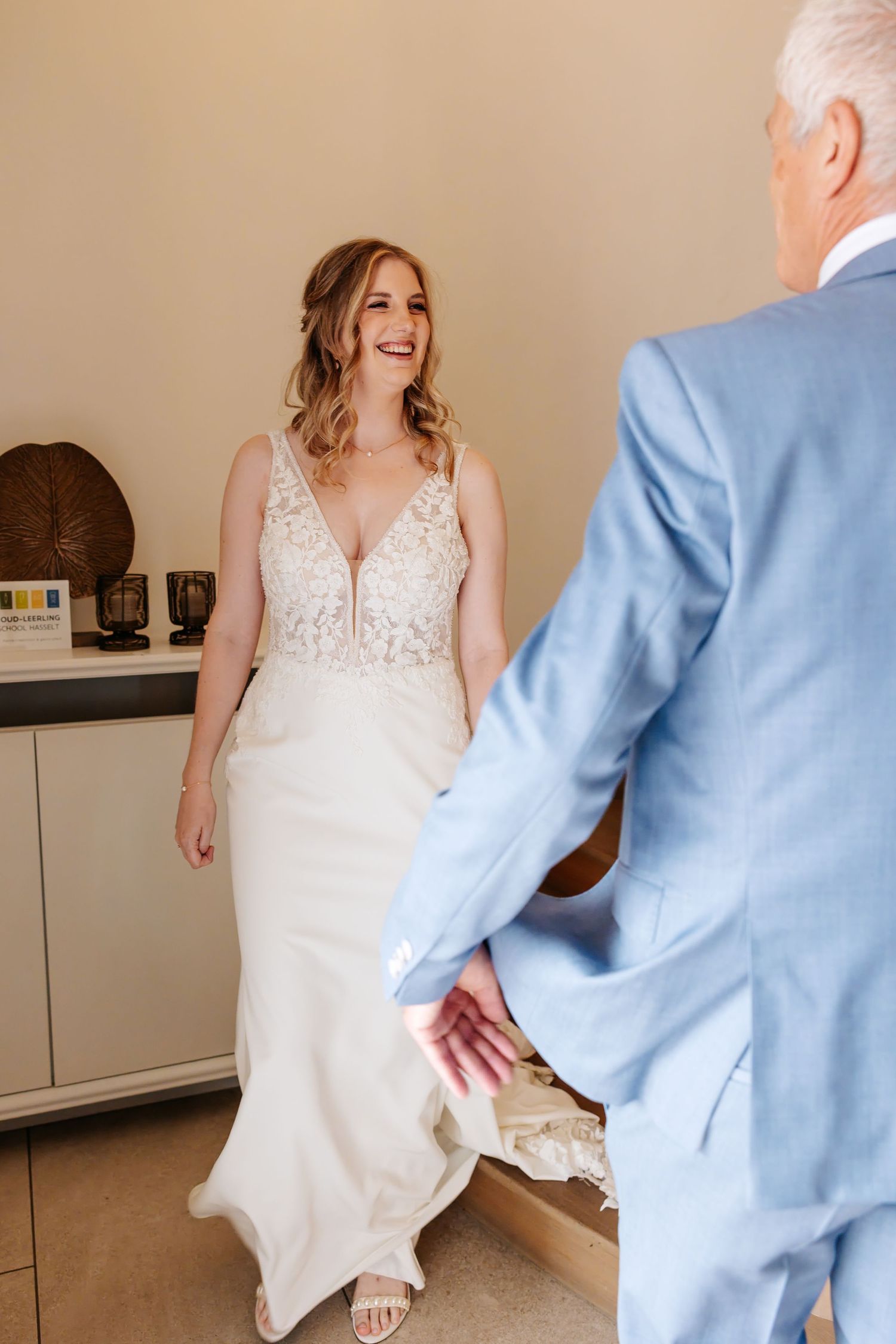 Smiling bride in lace wedding dress faces older man in blue suit during first look moment.