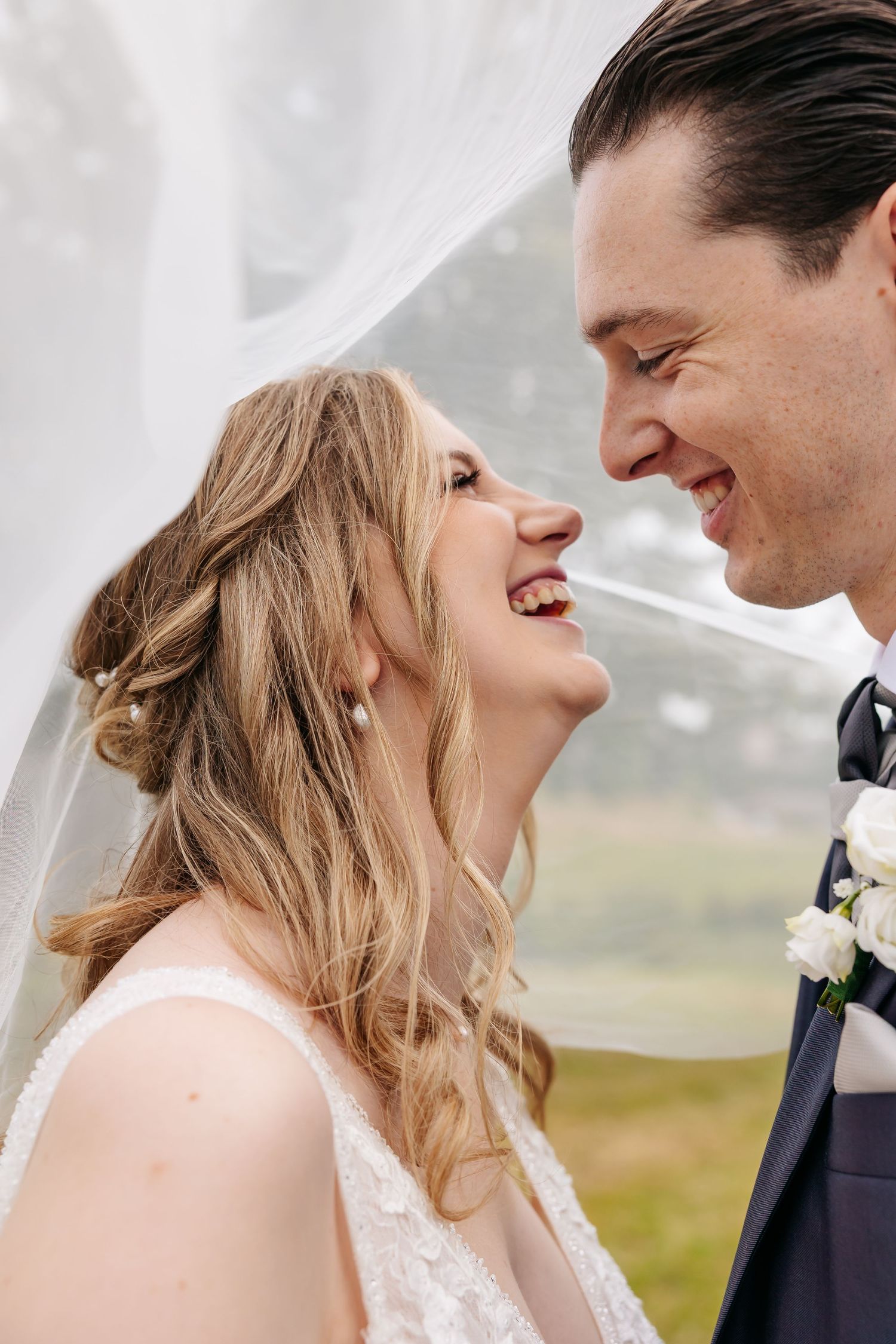 Close-up of bride and groom laughing together under a flowing veil, radiating pure wedding joy.