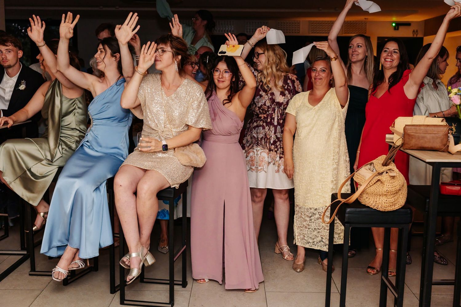 Excited wedding guests cheer with hands raised during a lively indoor wedding reception.