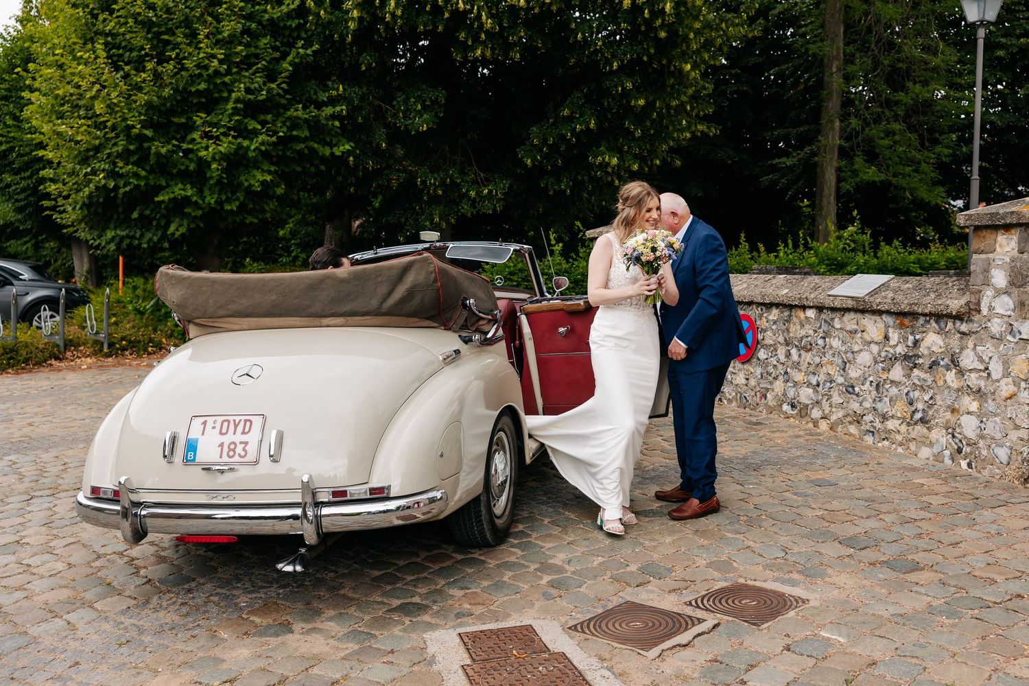 Bride in white gown and groom in blue suit share a kiss beside vintage cream Mercedes.