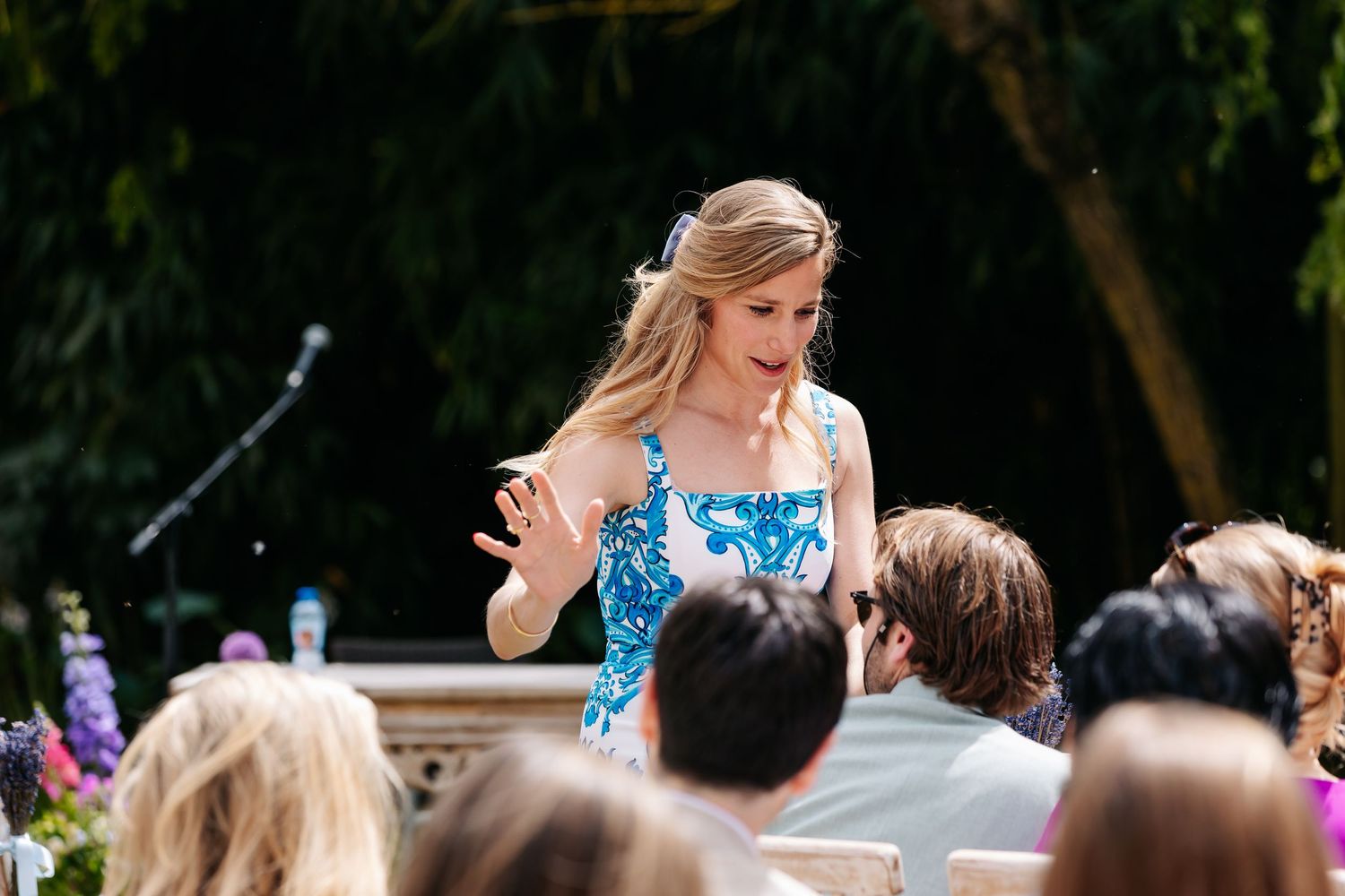 A woman in a blue floral dress gestures while speaking to seated wedding guests during an outdoor ceremony.