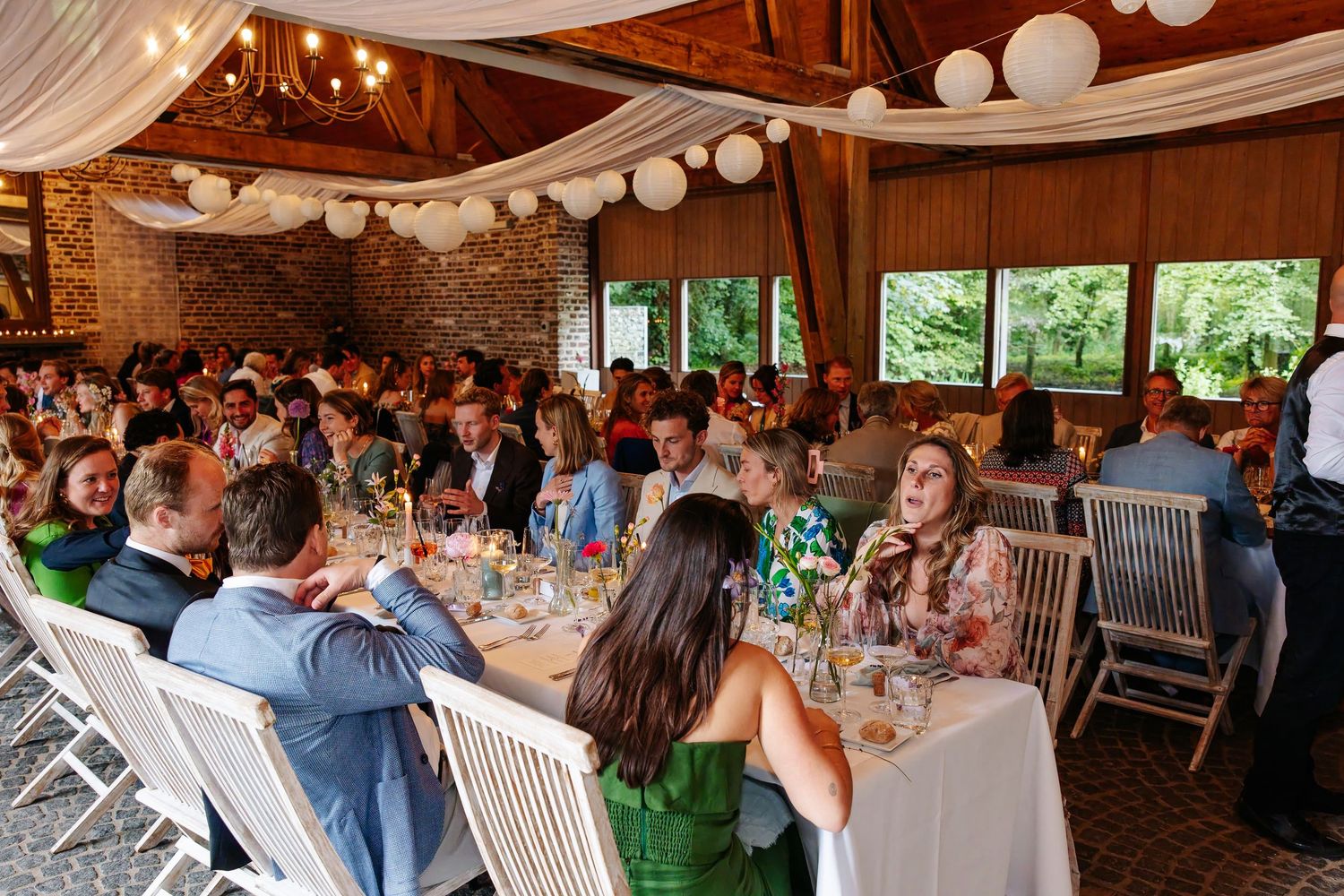 Wedding guests dine at round tables in a warmly lit rustic reception hall decorated with white lanterns.