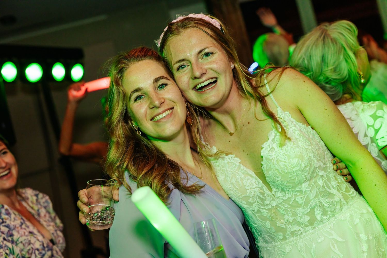 Two smiling women posing together at a wedding reception under green lights, one in a bridal gown.
