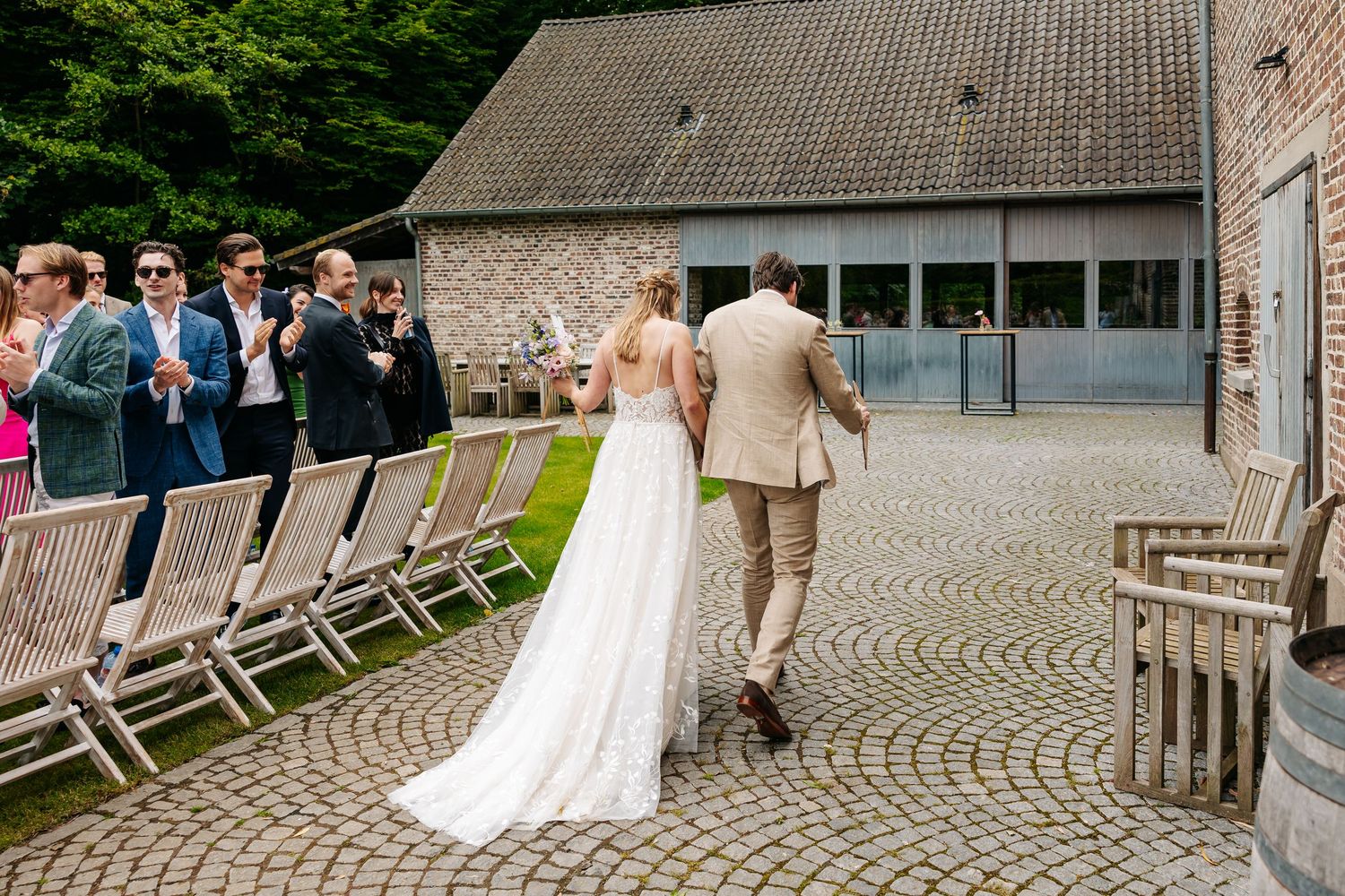 Bride in white gown and groom in beige suit walking toward brick venue entrance with seated wedding guests.