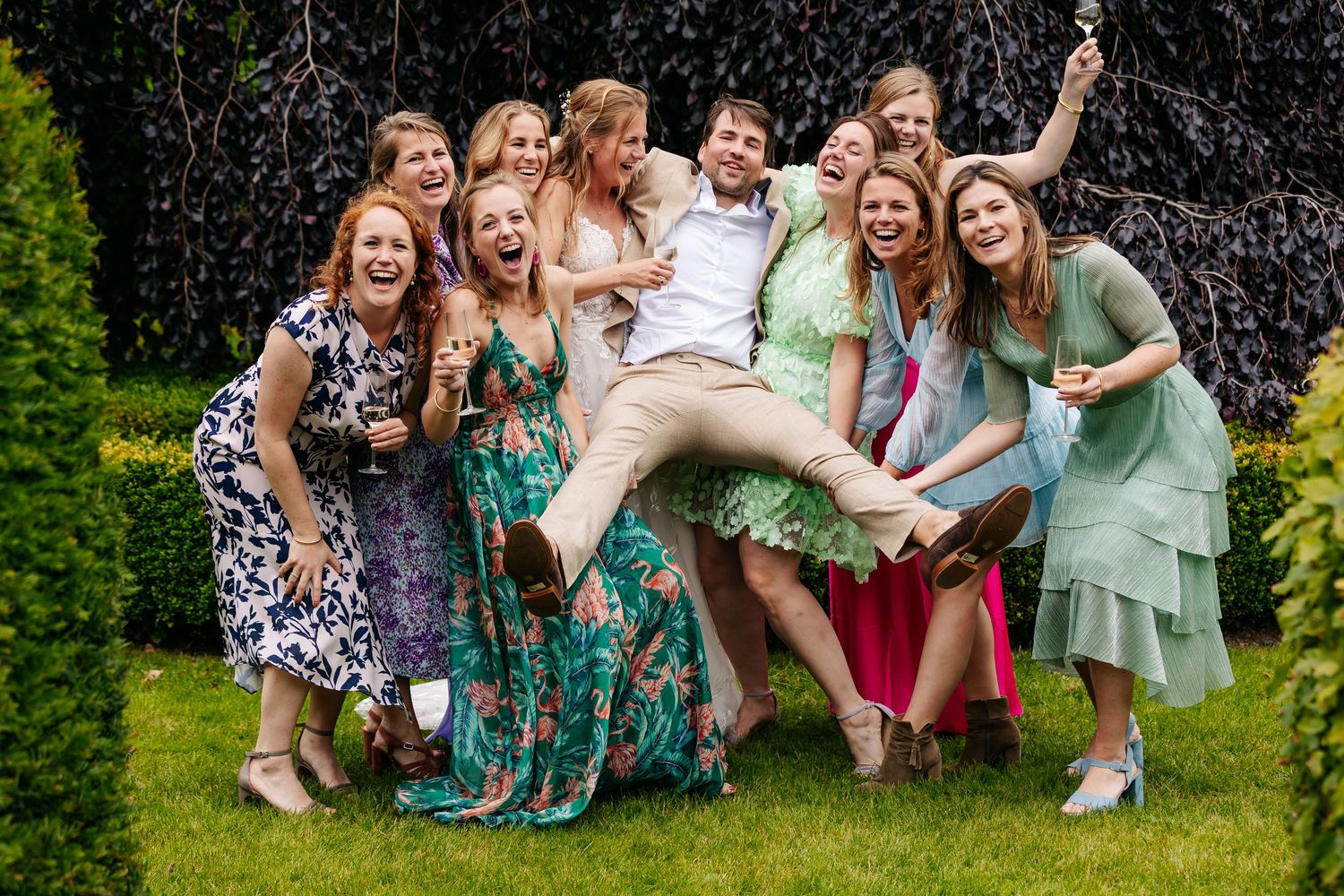Groom joyfully lifted by a group of smiling female wedding guests in a lush garden setting.