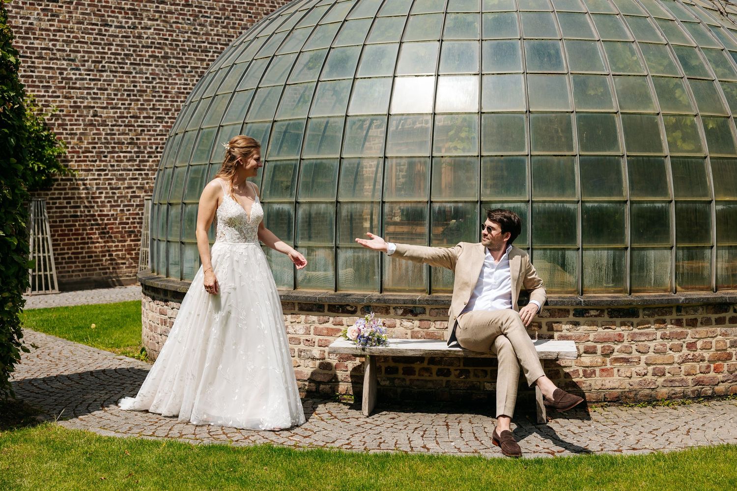A bride and groom posing playfully in front of a glass dome greenhouse on their wedding day.