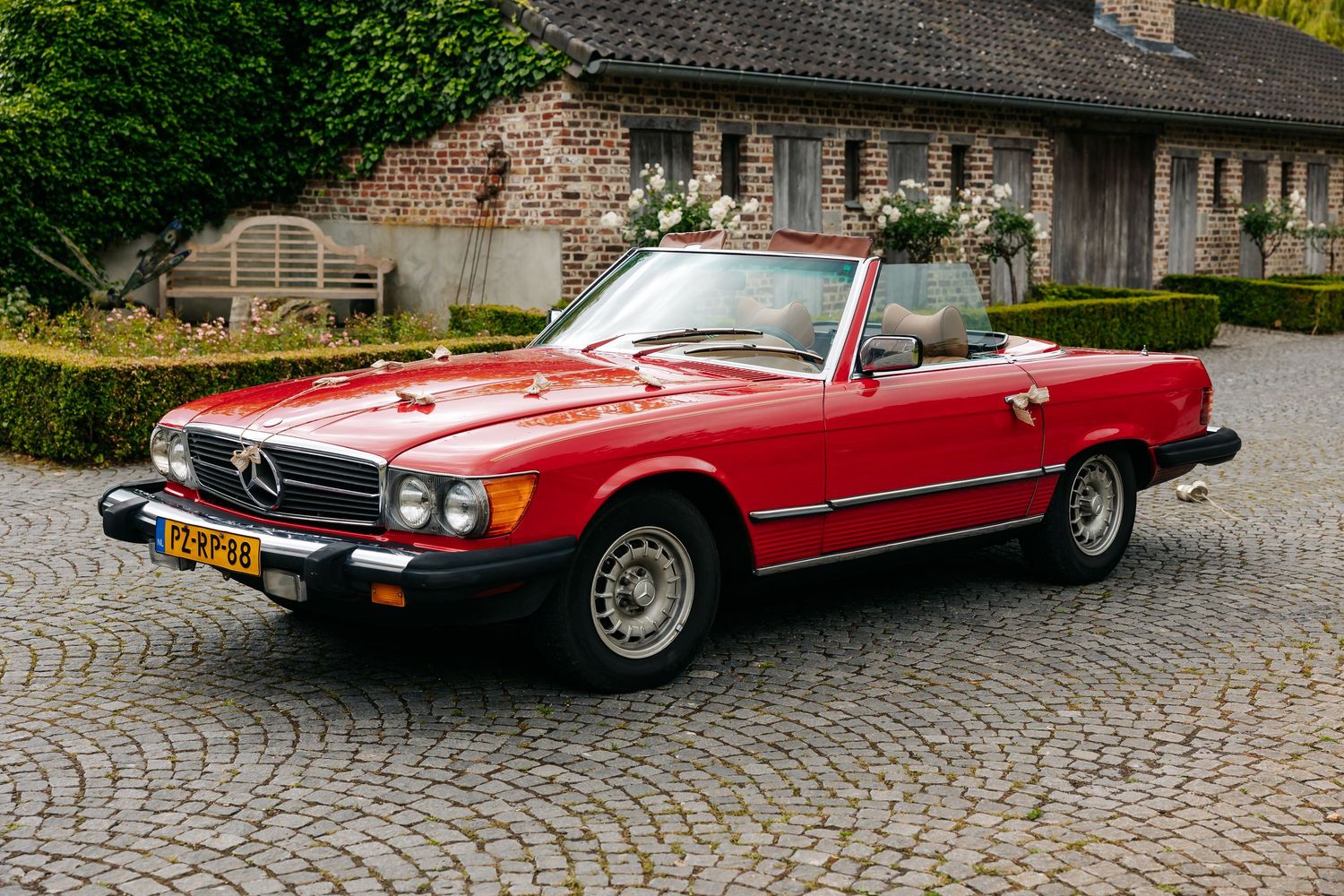 Classic red Mercedes-Benz 560SL convertible with floral wedding decoration on cobblestone driveway.
