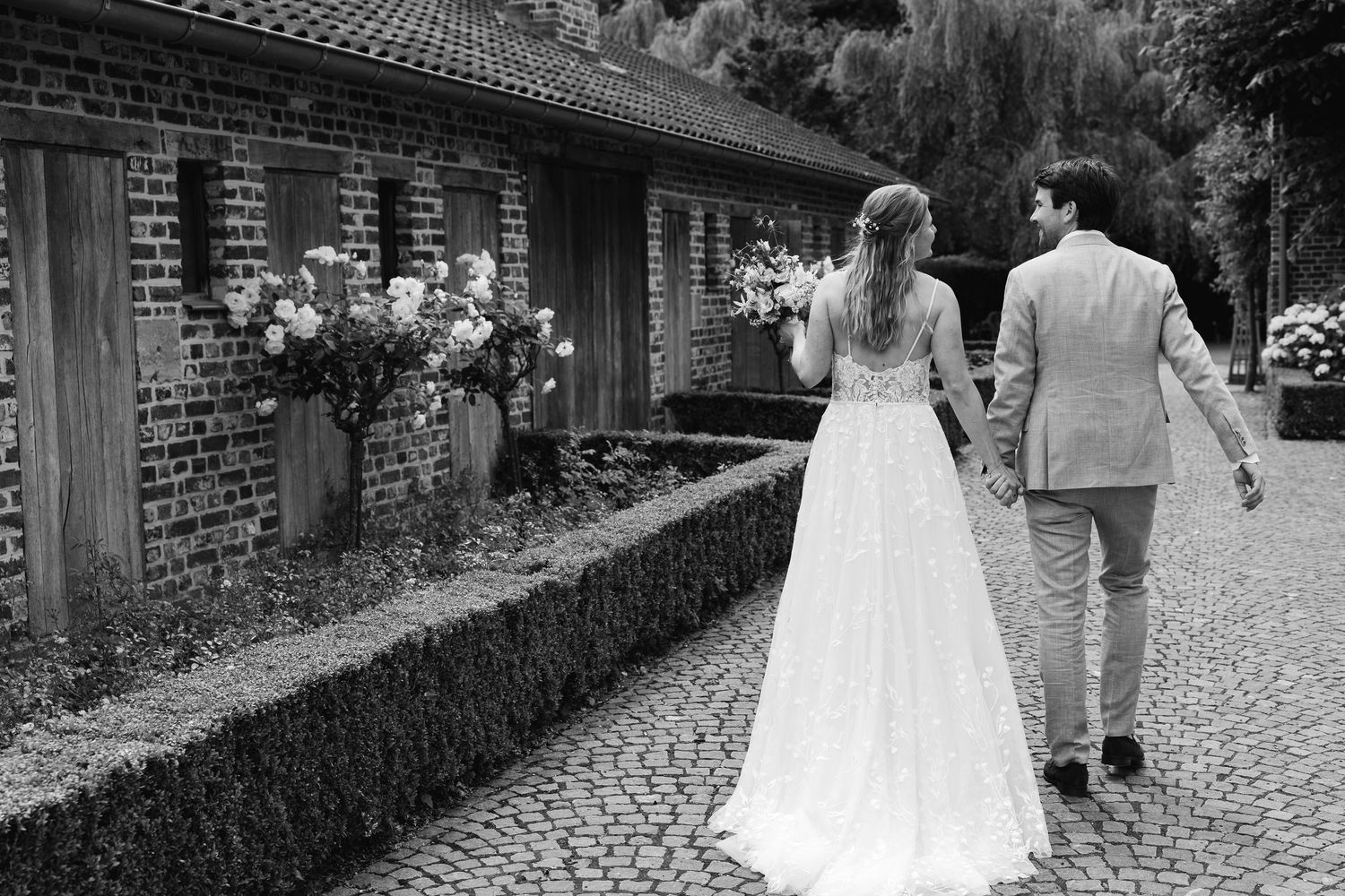 Black and white photo of bride and groom walking hand in hand past brick wall with floral arrangements.