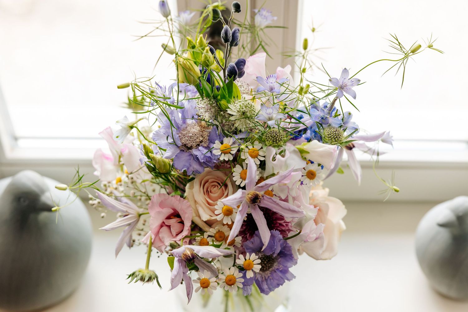 Elegant wildflower wedding bouquet with pink roses, purple blooms, and daisies arranged in a white vase by a window.