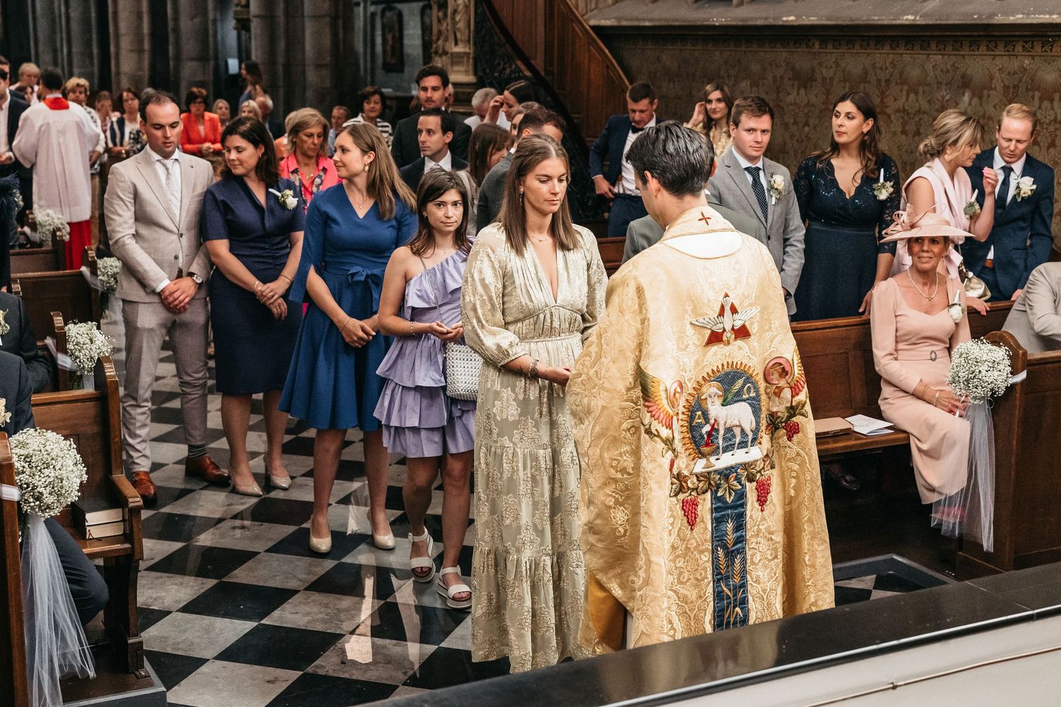 Priest in gold vestments greeting wedding guests in the aisle of a grand Gothic cathedral.