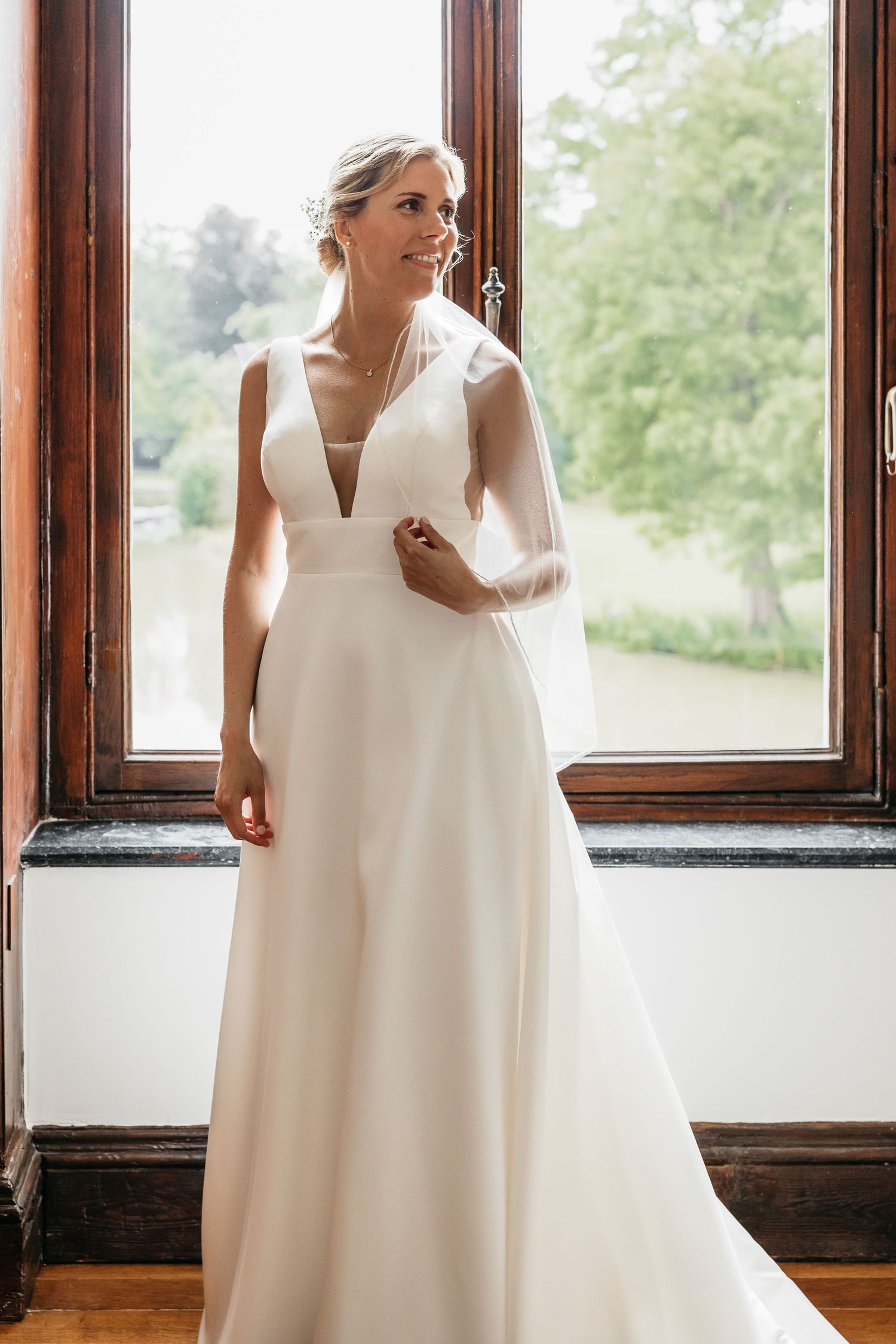Bride in elegant white deep V-neck wedding gown standing by wooden window with garden view.
